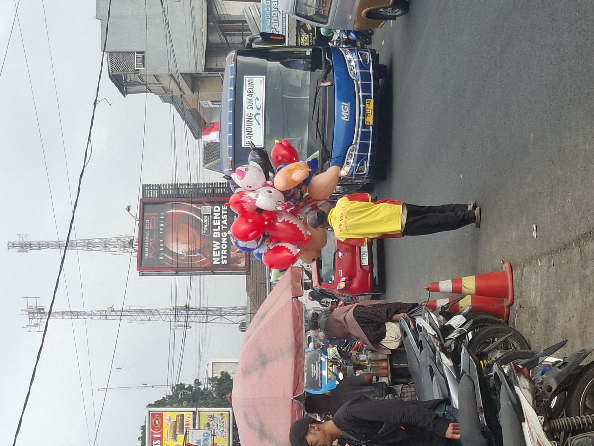 The image depicts a street scene in Gekbrong, Indonesia, with a blue bus prominently displayed. The bus has "BANDUNG-SUKABUMI" written on its front, indicating its route. A vendor is standing on the right side of the bus, holding a bunch of colorful balloons. The street is paved and appears to be a busy area with several motorcycles parked on the left side of the road. A red car is visible behind the vendor. Traffic cones are placed on the side of the road. In the background, two tall communication towers and billboards are visible. The sky is overcast.