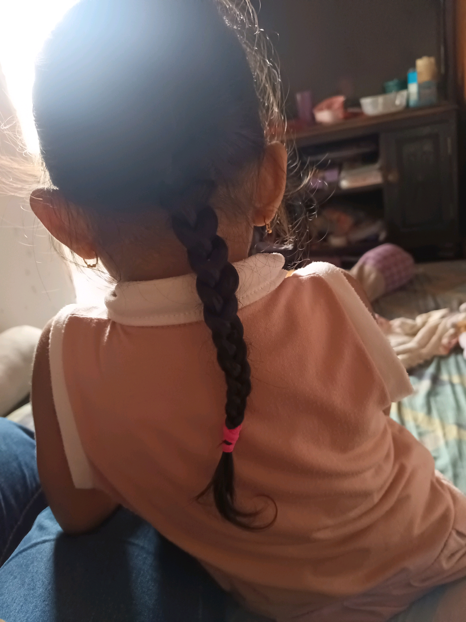 The image shows the back of a young child with dark hair braided and tied with a pink band. The child is wearing a peach-colored shirt with a white collar and appears to be sitting on someone's lap. In the background, a blurred cabinet with various items on it and a bed with patterned bedding are visible. The lighting suggests it is daytime, with sunlight streaming in from the left. The location is described as a residential area in South Jakarta, Indonesia.