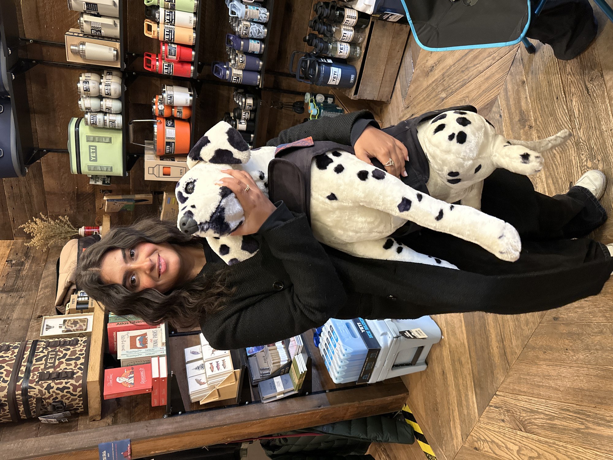 A woman with long, dark, curly hair and a warm smile is captured holding a large, black-and-white spotted plush dog, resembling a Dalmatian. She is wearing a dark, long-sleeved top or jacket and black pants, with rings visible on her right hand.
The scene is an indoor retail environment, likely an outdoor gear or lifestyle store, given the prominent display of numerous "YETI" brand products. These include various colored insulated bottles (some labeled "BOTTLE"), coolers, and "ICE BLOCK" ice packs, neatly arranged on wooden shelves.
In the background, behind the woman, more shelves hold a collection of books and notebooks. Visible book titles include "ROUGHING IT," "Discover wonder," "JOHN MUIR," and "NATIONAL PARKS," alongside "Atlas" and "FIELD NOTES" notebooks. A camouflage-patterned "YETI" bag is also seen. The store features a warm, rustic aesthetic with wooden walls and a wooden floor laid in a herringbone pattern.
The woman's pleasant expression suggests she is enjoying her time browsing or shopping. The abundance of outdoor-themed merchandise and literature indicates the store's focus. The location context provided places this store in the Village of Cold Spring, United States of America.