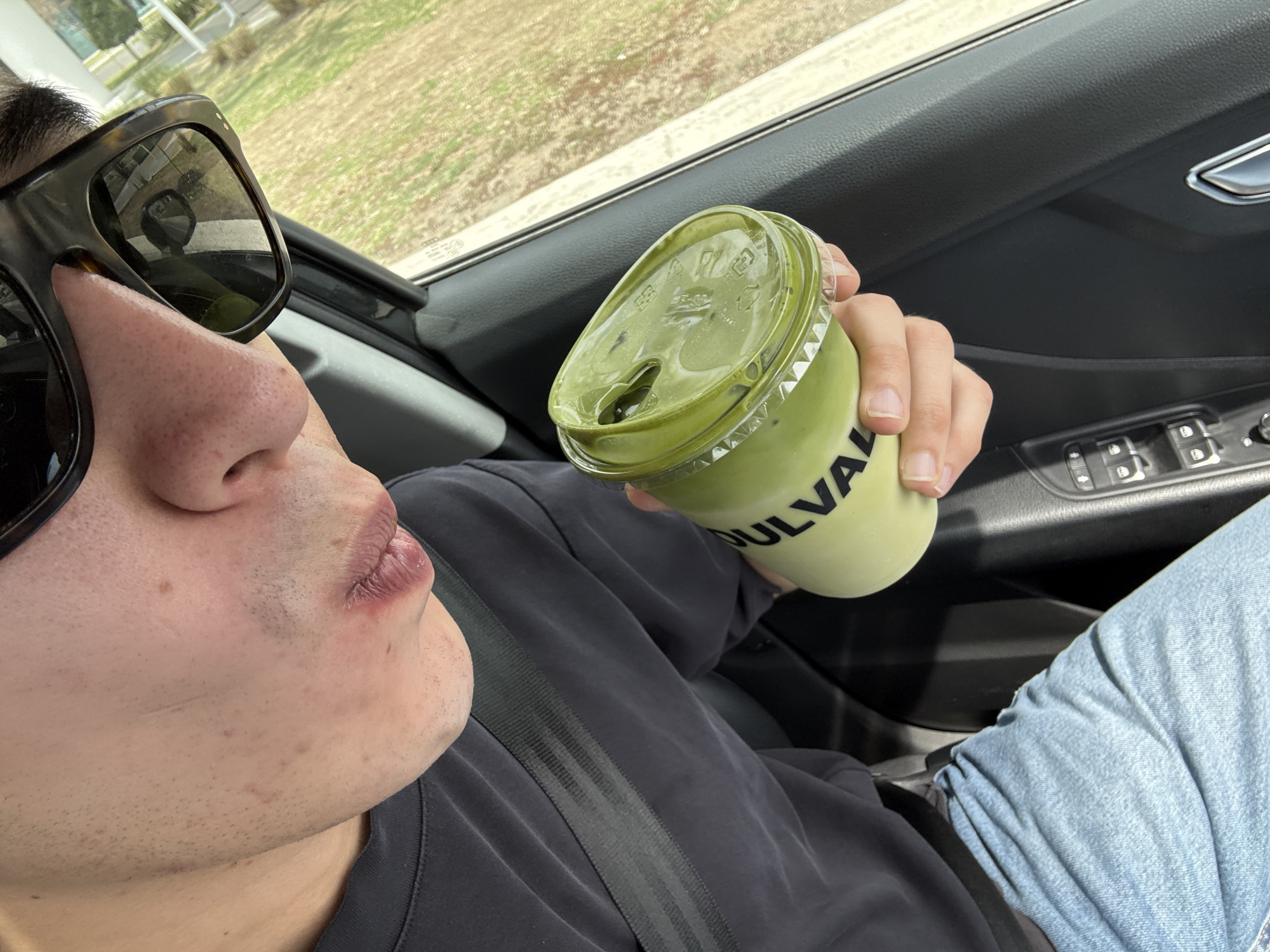A person wearing sunglasses and a seatbelt is sitting in a car, holding a green drink in a clear plastic cup with the brand name "SOULVA" visible on the side. The drink appears to be matcha-based, with a clear plastic lid. The car interior is visible, including the door panel with window and lock controls. The background shows a blurred view of an outdoor landscape, suggesting the car is in motion. The person seems to be on the verge of taking a sip of the drink. There is no information to confirm this photo was taken in the city of Puebla, Mexico.