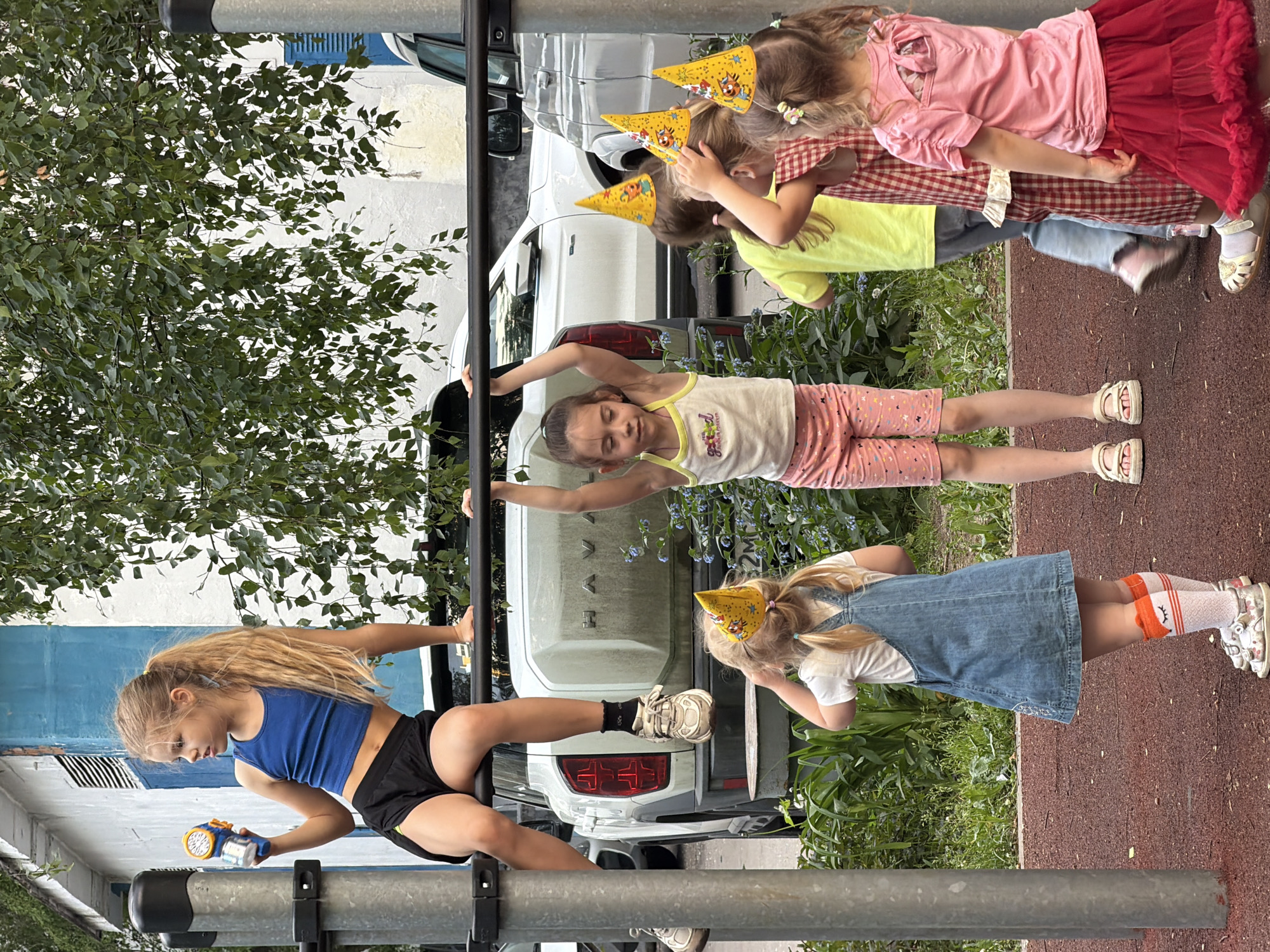The image depicts a group of children at what appears to be an outdoor playground or park area. One girl, wearing a blue athletic top and black shorts, is positioned on horizontal metal bars, seemingly engaged in a gymnastic exercise or play activity. She is holding an object resembling a toy or device in her hand.
Another girl is holding onto the bars with both hands, and is wearing a white tank top with pink shorts.
Three other girls, wearing party hats, stand behind the other two. Another girl in a denim dress and wearing patterned knee socks also seems to be watching.
Behind the children, part of a white SUV or car is visible, and there is greenery (trees and bushes) in the background. The ground appears to be red in the playground or park area.
Based on visual cues, there is not enough information to determine that this is Moscow, Russia.