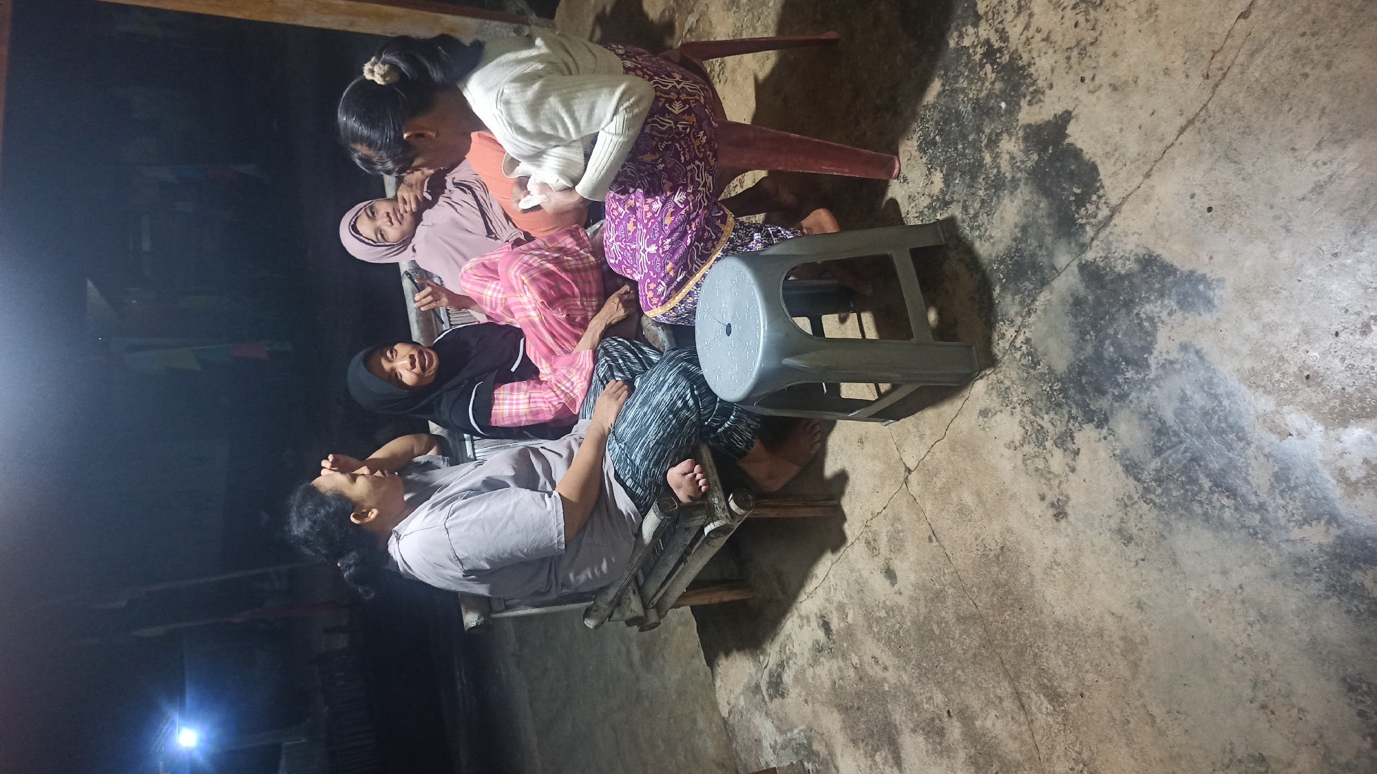 Four women are gathered outdoors, seated on a bamboo bench and a red plastic chair, against a concrete floor in what appears to be a rural setting in Tuba, Indonesia, likely at night. The lighting suggests artificial illumination, possibly from a nearby structure.
The woman on the far left, wearing a grey t-shirt and patterned pants, is looking down with her hand touching her forehead. Next to her, a woman in a black hijab and a pink and white checkered shirt is smiling and looking towards the right. In the middle, a woman wearing a light pink hijab and an orange top is looking forward with her hand near her chin. On the right, a woman with her hair pulled back in a bun is wearing a white sweater and a purple patterned skirt. She is holding a small object in her hands.
A grey plastic stool sits on the concrete floor in front of the group. The environment is dimly lit, with the background obscured by darkness, suggesting an outdoor space at night. The concrete floor shows signs of wear and cracking.