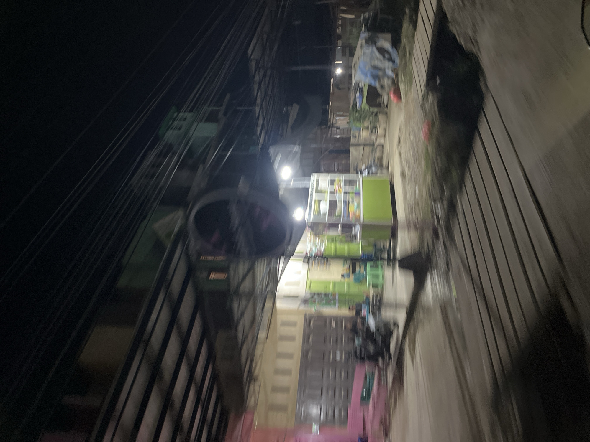 The image depicts a street scene at night in what is identified as Benuaraja, Indonesia. The perspective suggests the photograph was taken from a moving vehicle, resulting in motion blur throughout the image, particularly in the foreground and mid-ground.
In the mid-ground, several storefronts are visible. One building on the left has a pink facade and a large, corrugated metal roll-up door. Adjacent to it is a building with a cream-colored facade and several vertical window openings. Further to the right, there is a green structure which appears to be a food stall or small shop, with display shelves inside. There are also green doors visible on a building to the right of the pink building.
Several motorcycles are parked in front of the buildings. The ground appears to be a mix of paved surfaces and what looks like loose gravel or sand, with a wooden walkway or platform constructed over a dark area, possibly a ditch or water channel.
In the background, the scene is dominated by a dense network of overhead power lines and cables. Buildings are visible behind these wires, with some showing illuminated windows. The overall impression is of a densely populated urban or semi-urban area with active street-level commerce, illuminated by artificial lighting and the ambient glow of the night. A large, circular fan-like object is partially visible in the upper mid-ground, mounted above one of the structures.