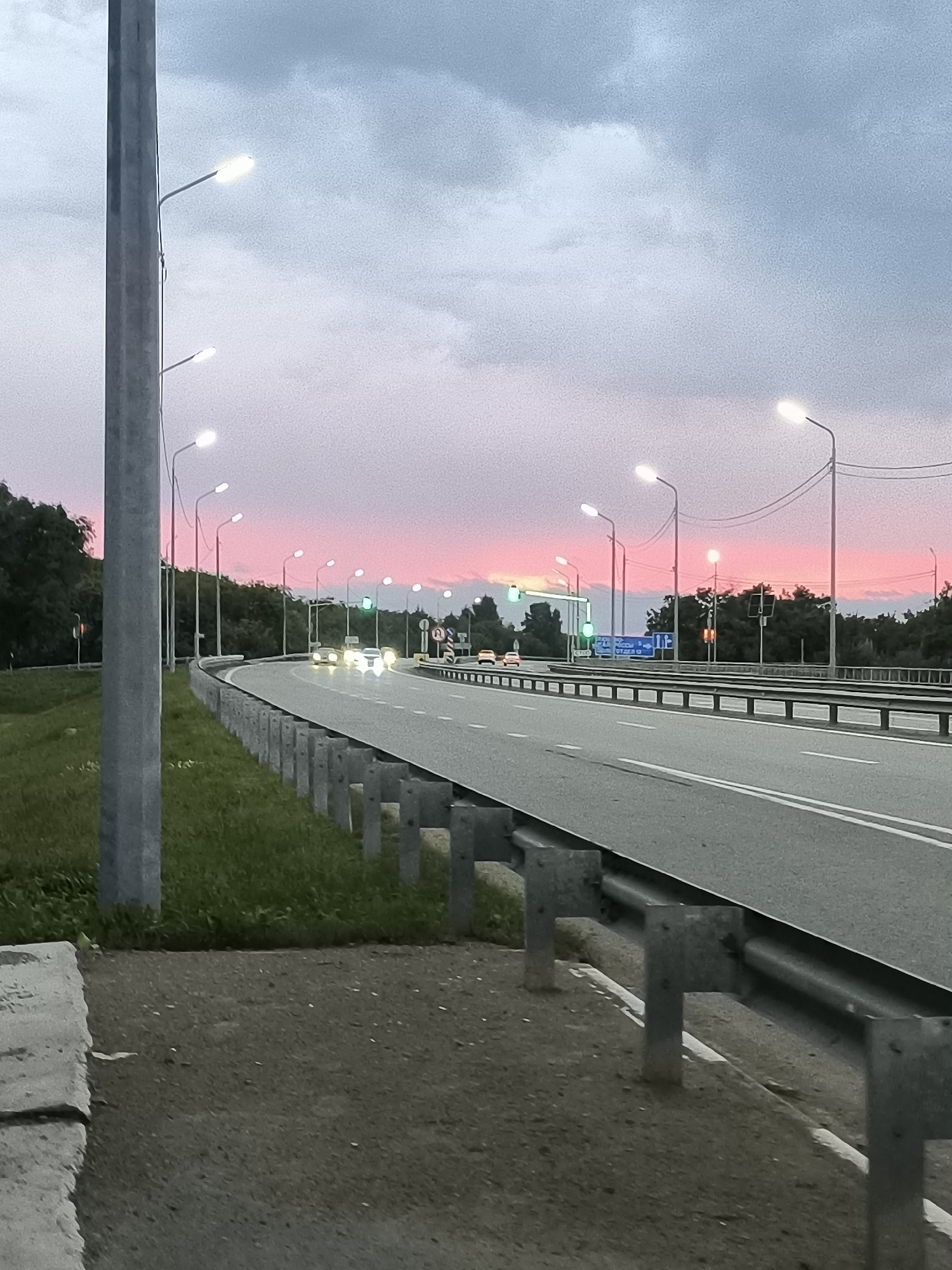 The image depicts a highway scene at dusk in Liubino-Malorossy, Russia. The sky displays a gradient of colors, transitioning from greyish-blue clouds at the top to a vibrant pink and orange sunset near the horizon. A concrete pillar with streetlights is prominent on the left side of the frame. The highway curves to the right, with multiple lanes visible. Several streetlights illuminate the road. Cars are driving on the highway, their headlights and taillights visible. A metal guardrail runs along the side of the road. On the left side of the highway, there is a grassy embankment with a concrete curb at the very bottom of the image. Trees line the background on both sides of the highway. Traffic signs are visible in the distance. The overall scene suggests a transitional time of day, with artificial lights beginning to dominate as natural light fades.