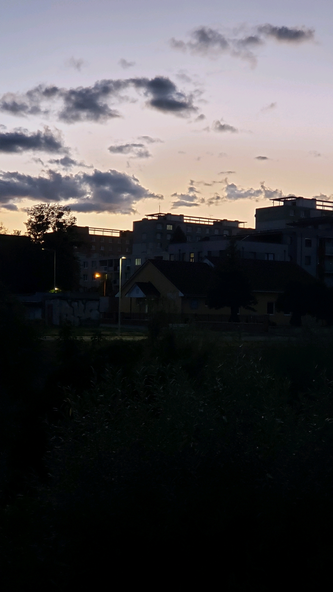 The image depicts a twilight scene in a residential area of Prešov, Slovakia. In the foreground, dark, out-of-focus foliage obscures the lower portion of the frame. Behind this, a low-lying building with a yellow facade and a dark roof is visible, with a small porch or entryway. To the left of this building, a modern lamppost illuminates the scene with a bright, white light. Further in the background, several multi-story apartment buildings are silhouetted against the gradient sky, which transitions from a pale purple and pink near the horizon to a darker blue at the top, with scattered grey clouds. The windows of the apartment buildings show faint internal lights, suggesting the time is early evening or late twilight. A single, less modern lamppost with an orange glow is visible between two of the apartment buildings. A large tree with dark foliage stands to the left of the apartment blocks, partially obscuring them. The overall atmosphere is quiet and subdued, indicative of a typical evening in a suburban or residential district.