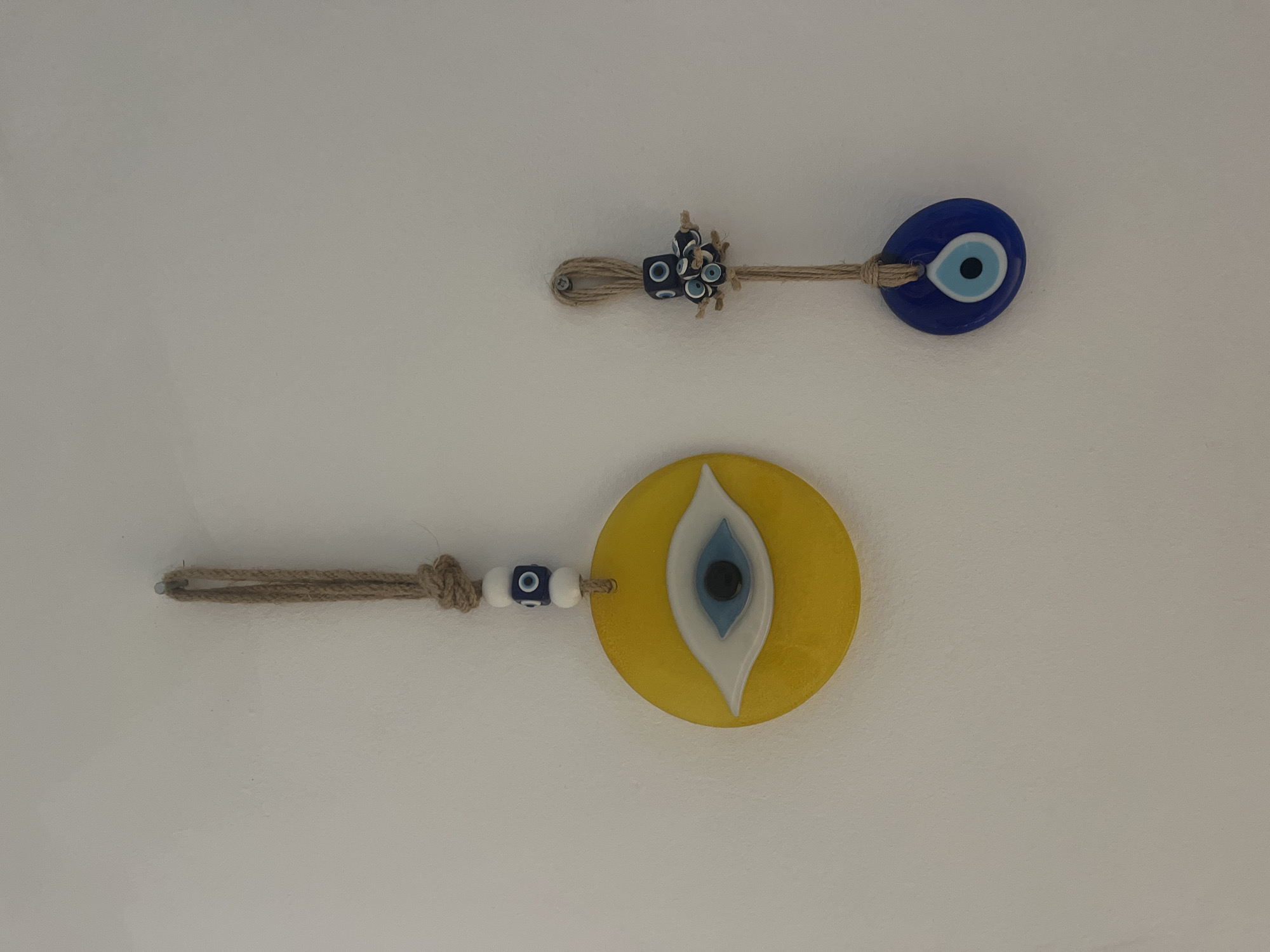 The image displays two decorative "evil eye" charms hanging against a plain, light-colored wall, likely indoors.
The upper charm is smaller, featuring a dark blue, oval-shaped evil eye bead. This bead is attached to a loop made of twisted natural fiber rope, possibly jute. A small cluster of miniature blue and white beads is intricately tied into the rope near the main evil eye. The rope's looped end is secured to the wall by a small, visible metal hook or nail.
The lower charm is larger and circular, rendered in a vibrant yellow. Its central design depicts a stylized eye with distinct layers of white, light blue, and a central black pupil. This charm is also suspended by a similar twisted natural fiber rope. Two small blue and white patterned beads are threaded onto this rope before it connects to the large yellow evil eye. The rope forms a loop at its far end, prepared for hanging, though the exact attachment point to the wall is not fully visible.
The overall scene is static, showcasing these decorative items. There is no activity or event taking place. The charms, known as Nazar amulets or evil eyes, are widely recognized cultural symbols in Mediterranean regions, including Spain, which aligns with the provided location context of Barcelona. No visible text is present in the image.