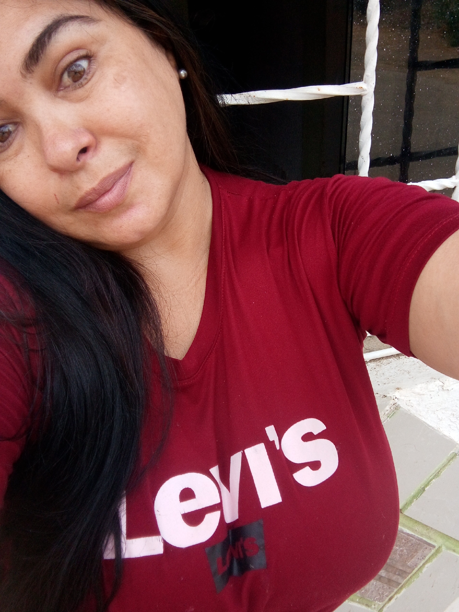 A person is posing for a selfie outdoors. The person has dark hair, light brown eyes, and is wearing a maroon Levi's T-shirt. Behind the person is a white metal window frame and a dark wall. The exact location is unknown.