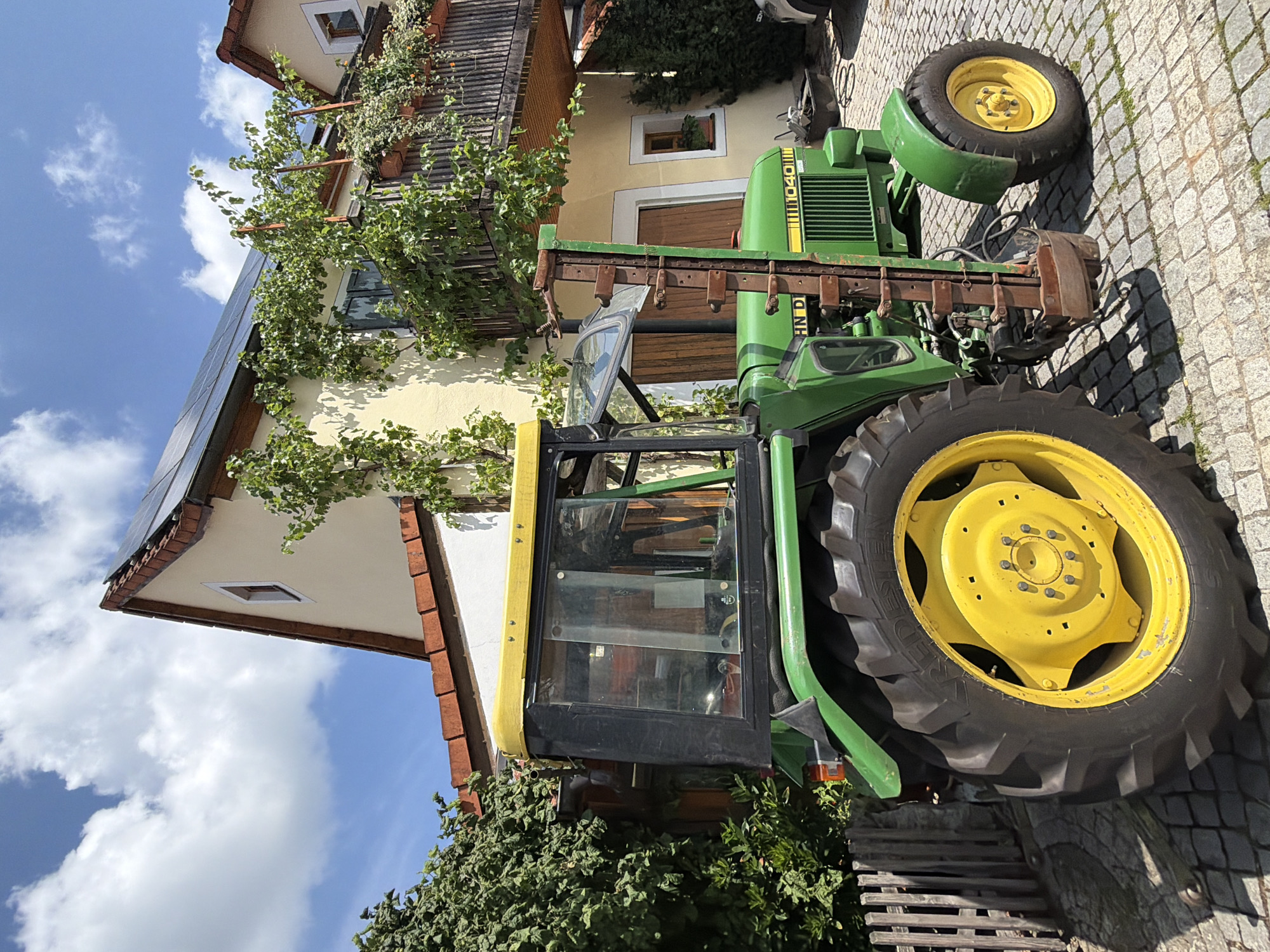 A John Deere 1040 tractor is parked on a cobblestone driveway in front of a yellow house with solar panels on the roof in Ebermannstadt, Germany. The tractor is green and yellow, with large rear tires and a smaller front tire. A green hedge is to the left of the tractor, and a wooden fence is behind it. The house has a balcony with plants and a vine growing on the side. The sky is blue with white clouds.
