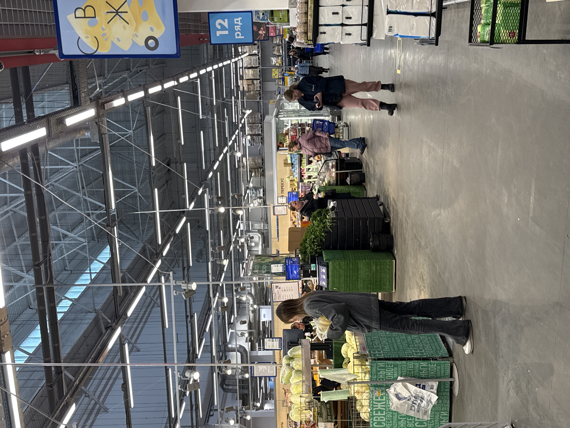 The image shows the interior of a supermarket, potentially in Izhevsk, Russia, given the Cyrillic text on the signs. The perspective is from an aisle, looking down its length. The building has a high, exposed metal roof structure with rows of fluorescent lights illuminating the scene. The aisle features a variety of products, including cabbages in wire baskets near the foreground, indicated by the sign "СВЕЖЕЕ..." ("FRESH...") in Cyrillic, with green product boxes underneath the cabbages. There are signs in the distance and directly above the cabbages, including "И ПЕРЕКУС" ("AND SNACKS") and a blue sign with a cheese graphic labelled "В Ж". Above the cabbages is a sign that says "ТОВАРОВ".
Several people are shopping in the aisle. Closest to the camera, a woman in a grey sweater is examining the cabbages. Further down the aisle, other shoppers are visible, including a woman looking at her phone and another pushing a shopping cart. In the upper right corner, stacked boxes of goods are visible. The floor is a plain grey concrete. The general atmosphere suggests a large, busy supermarket environment.