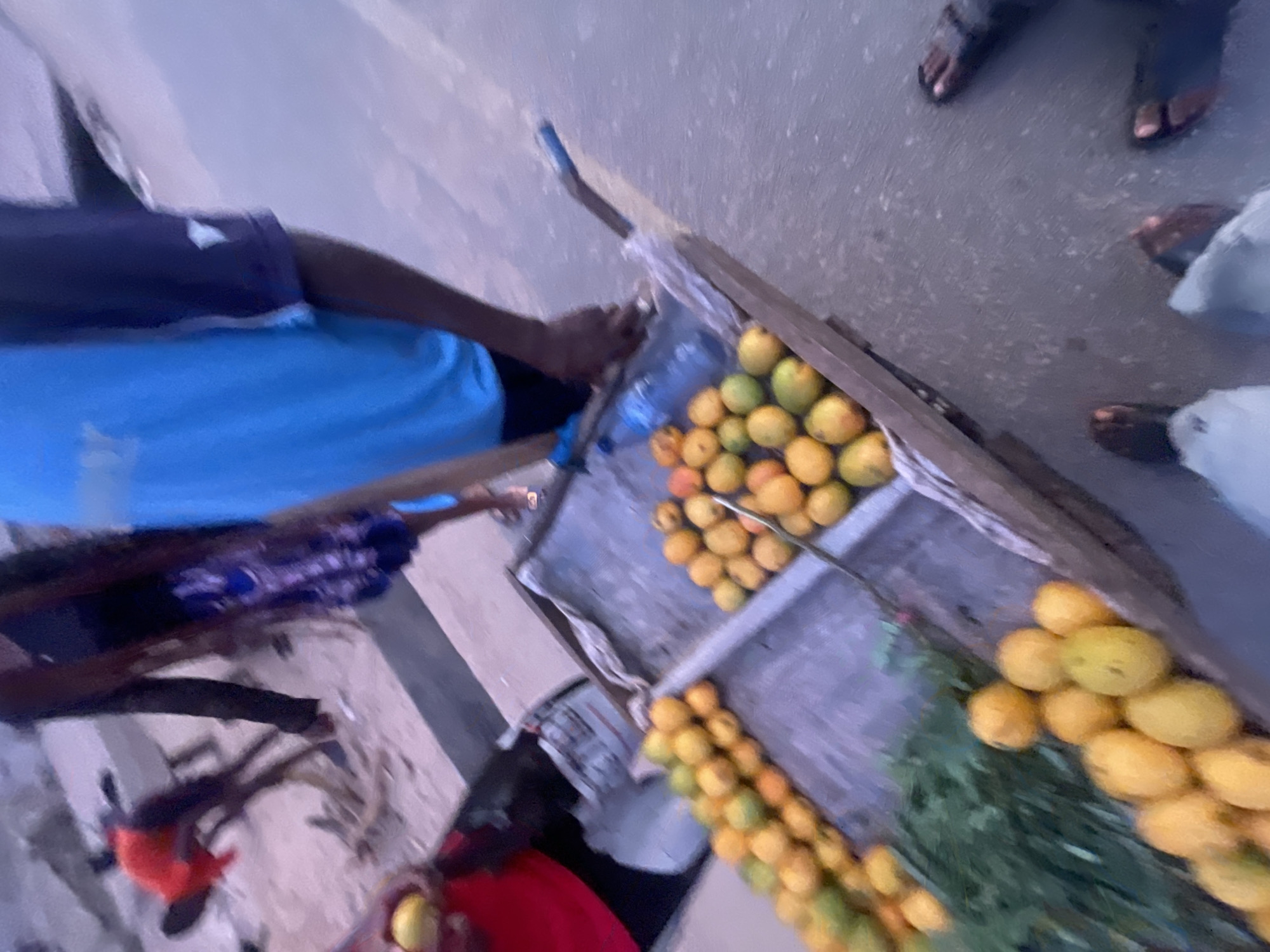 The image depicts a street vendor in Lokoja, Nigeria. He is selling fruit from a cart, including mangoes and oranges. He is wearing a blue shirt and black shorts. There are customers standing near the cart.