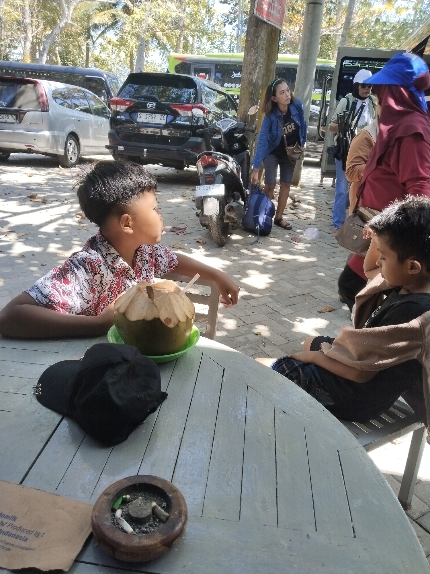 In Prigi, Indonesia, a group of people are gathered near a street. In the foreground, two young boys are seated at a table. The boy on the left, wearing a patterned shirt, looks to his right, while the boy on the right, wearing a black shirt, looks down at his lap. A whole coconut with a straw is placed on a green dish on the table in front of the boy on the left. A black baseball cap and an ashtray with cigarette butts are also on the table. In the background, several vehicles, including cars and buses, are parked. A woman in a blue jacket and shorts is standing near a motorcycle and holding a blue bag. Another woman, wearing a blue headscarf and a red jacket, is standing and looking towards the left. The ground is paved with light-colored bricks, and the scene is shaded by trees.