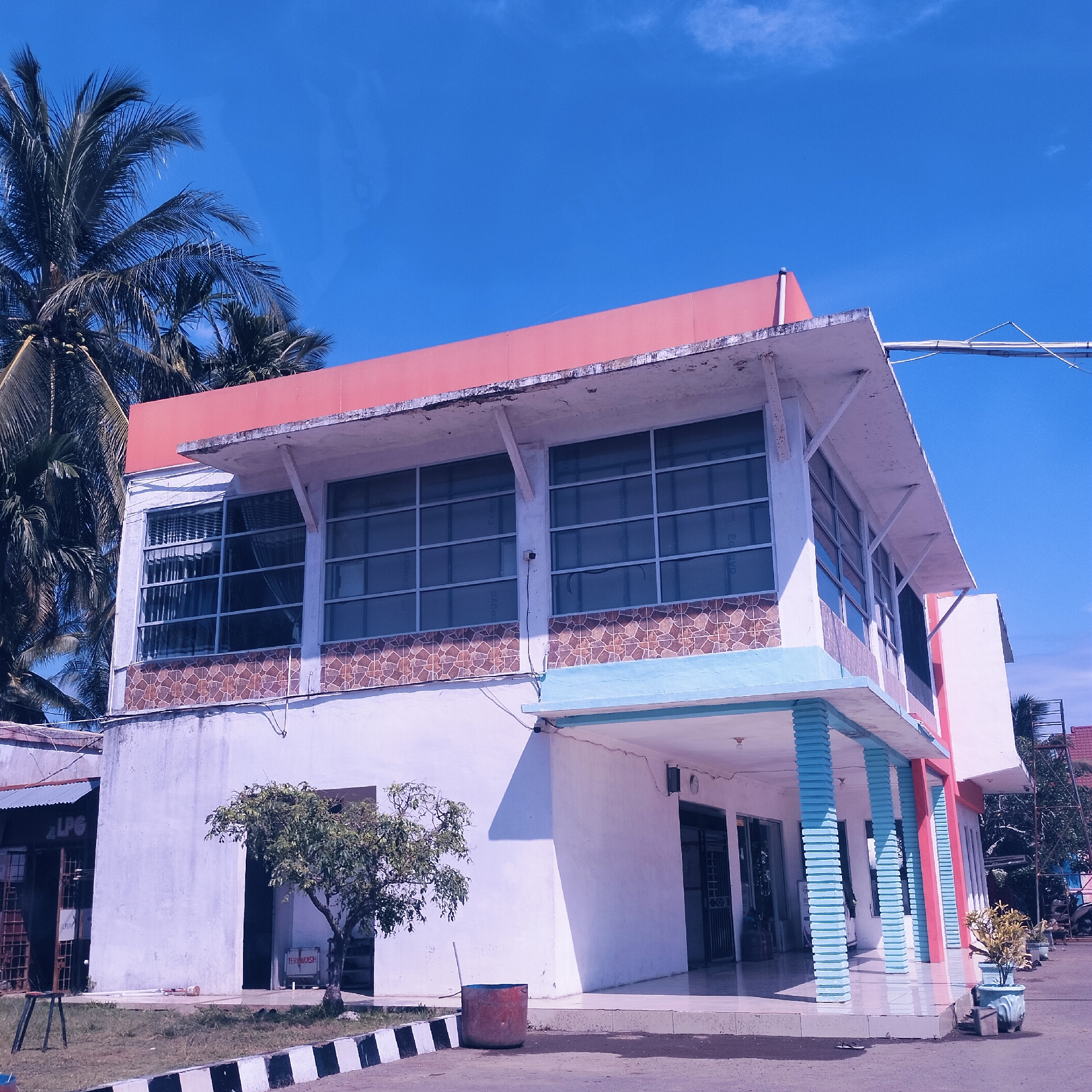 The image displays a two-story building under a clear, bright blue sky, typical of a sunny day. The main structure features white walls, with a prominent red roofline or trim on its upper level. The second floor is characterized by large, grid-patterned windows that reflect the sky, and a decorative band of brown and orange tessellated tiles below them.
On the ground level, a covered entrance area is supported by multiple light blue, horizontally ribbed pillars, with additional red pillars visible further back on the right. A small, leafy green tree stands in front of the building, and a red barrel sits on the paved ground nearby. To the far left, a more rustic, single-story structure with a corrugated metal roof is partially visible, with a sign reading "LPG." Inside an entrance on the main building's lower floor, a sign with the Indonesian word "TERIMAKASIH" ("Thank you") is displayed.
Several large palm trees with green fronds are prominent on the left side of the frame, reaching towards the almost cloudless sky. The foreground includes a paved area and a black and white striped curb. No people or specific activities are visible in the scene, which appears to be set during daytime given the strong sunlight and blue sky. The presence of palm trees and Indonesian text suggests a tropical location, consistent with Cotpuuk, Indonesia.