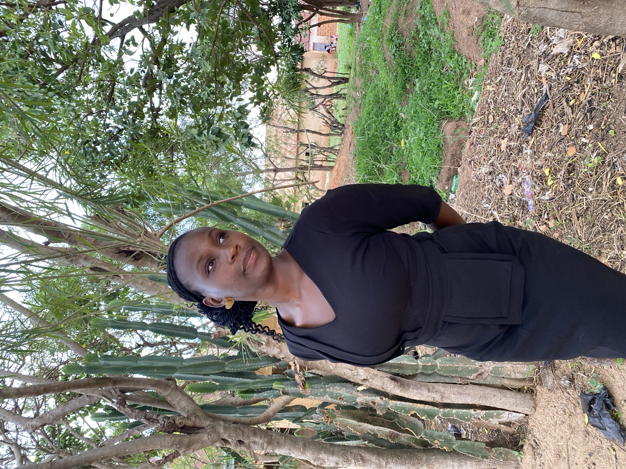 The image shows a woman in a black dress standing outdoors. She is positioned in front of a tree or bush with long, green stems resembling cacti. The background includes other trees and some green vegetation. In the distance, there is a building. There is a variety of dried leaves on the ground. Based on the prompt information, the location is Kuru Dungus, Nigeria.