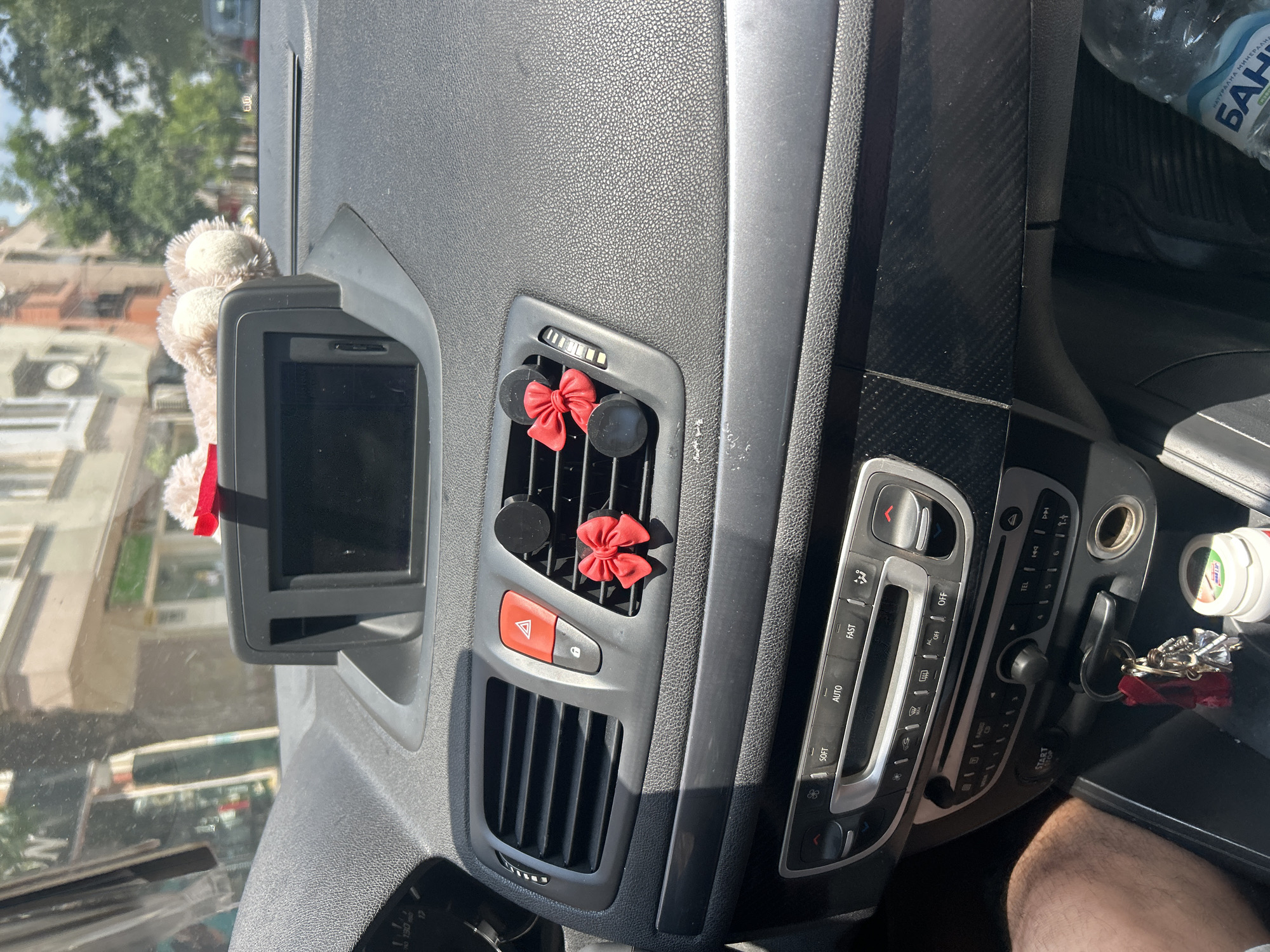 The image shows the interior of a car parked on a street in Plovdiv, Bulgaria, during daytime. The car's dashboard is visible, featuring a central display screen, air vents with red bows attached, hazard lights button, and various car controls and buttons. A teddy bear is partially visible on top of the dashboard. The exterior view through the windshield reveals buildings and trees, characteristic of an urban environment. A bottle of water and keys are visible in the lower part of the image.