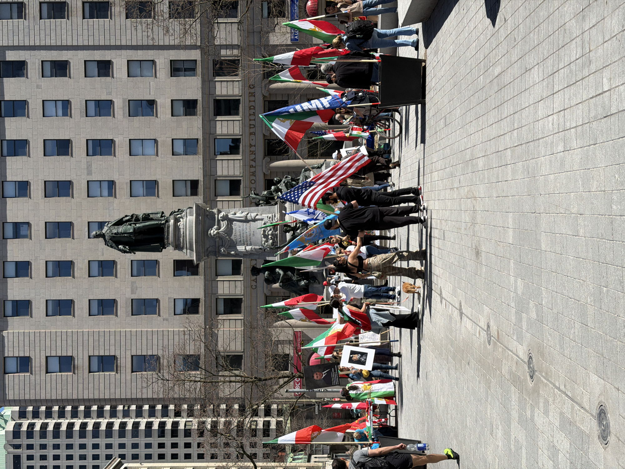 The image depicts a gathering of people in Montreal, Canada, identifiable by the "COMPANY-BUILDING" sign on the side of the building, the statue in the foreground, and the Iranian flags held by several people in the crowd. A mix of people are standing and holding flags. Some are walking or running on the right side of the image. The scene takes place on a paved area near a building and a statue in Montreal. An American flag and other flags are also visible among the participants.