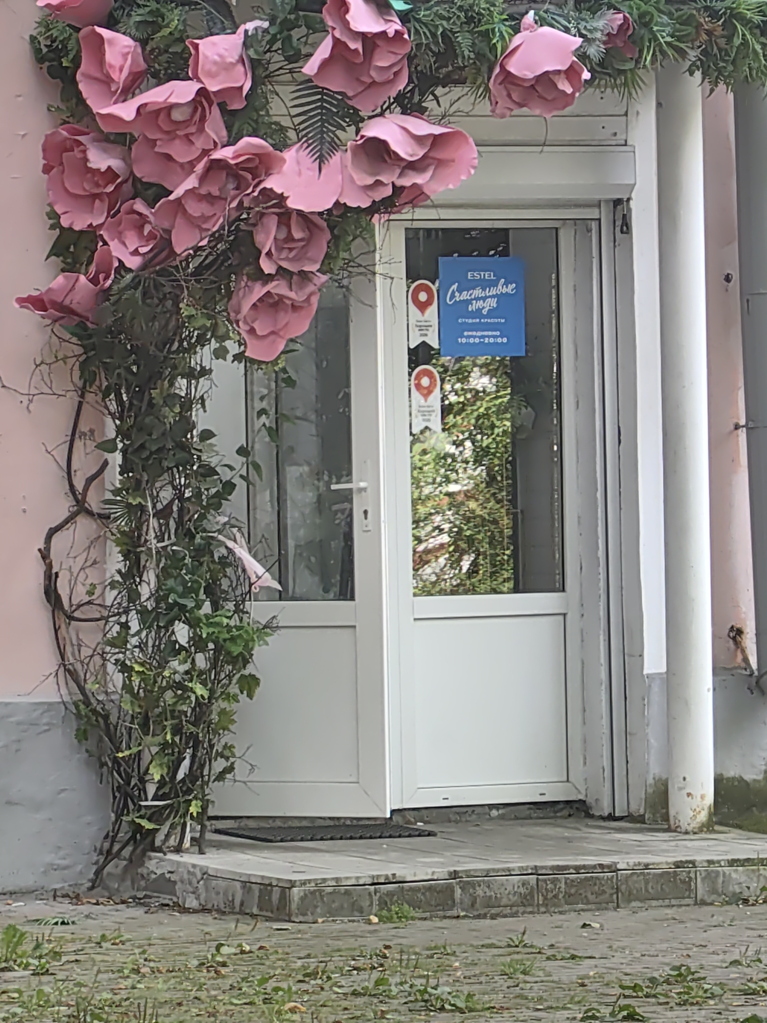 This is an outdoor, eye-level medium shot of a storefront entrance decorated with large, artificial pink roses and greenery. The storefront features a white door with a glass pane and a white metal rolling shutter above it. A blue sign is affixed to the glass door with Russian text that reads "ESTEL Счастливые люди" (ESTEL Happy People) and "СТУДИЯ КРАСОТЫ" (Beauty Studio), along with operating hours. The entrance is raised from the sidewalk, with stone steps leading up to it. The surrounding environment includes a pink wall on the left, a white pillar on the right, and a cobblestone walkway with scattered leaves and grass in the foreground. The image does not contain any identifiable people or actions. The location is Tver, Russia.
