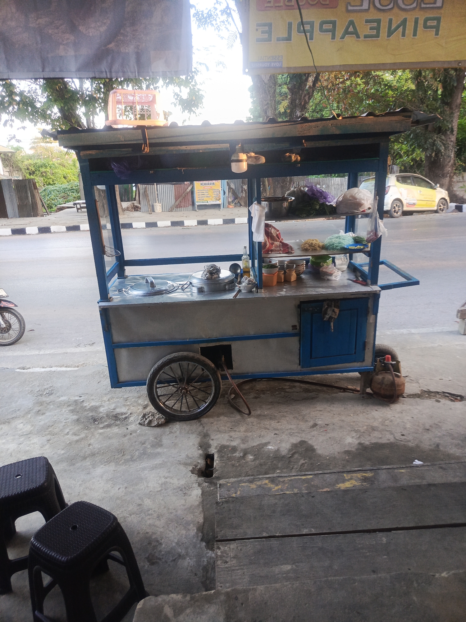 A street food cart is parked on a sidewalk in Kendari, Indonesia. The cart is blue and silver, with a corrugated metal roof. It is stocked with various food items, including vegetables, noodles, and what appear to be pots of soup or stew. A propane tank is attached to the cart, suggesting it is used for cooking. Two plastic stools are placed in front of the cart, and a motorcycle is parked to the left. In the background, a yellow car is visible on the street, and trees line the roadside. A banner advertising "PINEAPPLE JUICE" is hanging above the cart. The scene appears to be during daylight hours, with soft, diffused lighting.