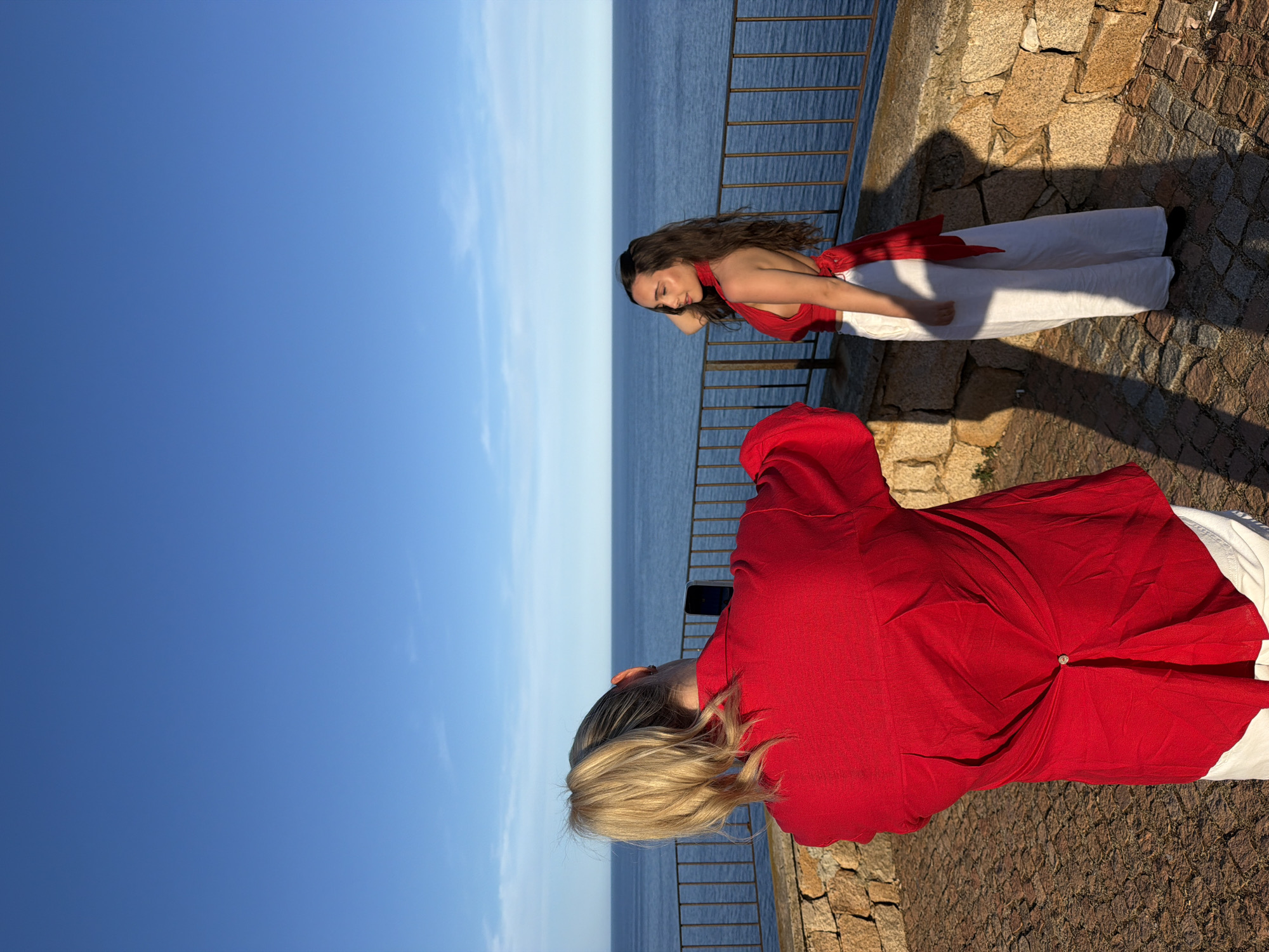 In Santa Maria Navarrese, Italy, two women are captured in a photograph on a cobblestone terrace overlooking the sea. A woman with blonde hair pulled into a ponytail, wearing a red shirt and white pants, is taking a picture with her smartphone. Facing away from the camera, she is positioned in the foreground. To her right and slightly further back, a woman with long, dark curly hair is posing for the photo. She is wearing a red halter-neck top with a tie detail in the back and flowing white pants. The background features a clear blue sky meeting the calm, blue sea, separated by a metal railing. The stone wall of the terrace is visible in the lower part of the image. The lighting suggests it is late afternoon, casting distinct shadows on the ground.