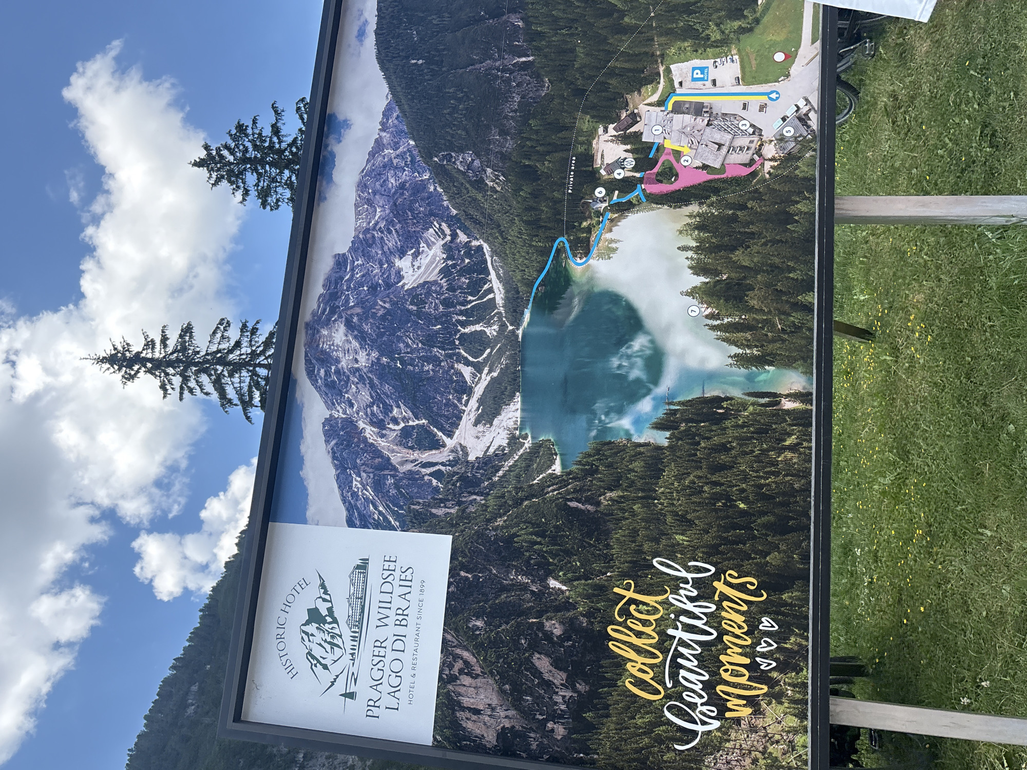 This is a photograph of a large sign displaying a scenic mountain and lake landscape with a superimposed map. The sign is positioned outdoors, with a grassy area in the foreground and trees and a cloudy sky in the background. The sign features a logo for "HISTORIC HOTEL PRAGSER WILDSEE LAGO DI BRAIES HOTEL & RESTAURANT SINCE 1899". The main image on the sign shows a large, clear lake reflecting snow-capped mountains and surrounded by dense evergreen forests. A map is overlaid on the right side of the image, indicating a hotel complex with numbered points of interest, pathways, and parking areas. Text on the lower left of the sign reads "collect beautiful moments" in a decorative script, with small heart symbols below. The sign is supported by metal frames and appears to be standing on an outdoor field.