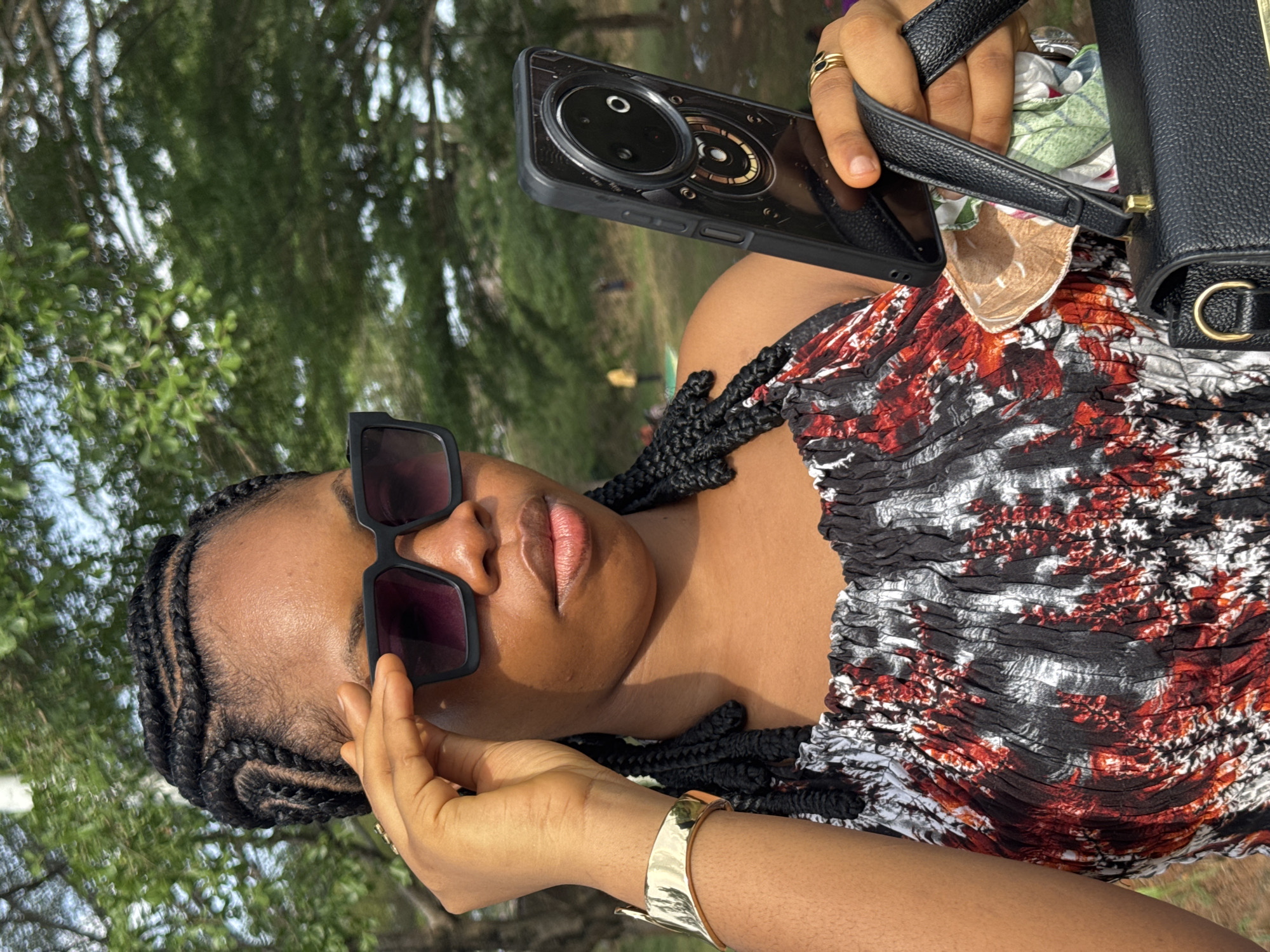 A woman is holding a black mobile phone in her right hand. She is wearing black sunglasses, a black and red patterned top, and a gold bracelet. Her hair is styled in braids. The background shows green trees, suggesting an outdoor setting. The image was taken in Ihiagwa, Nigeria.