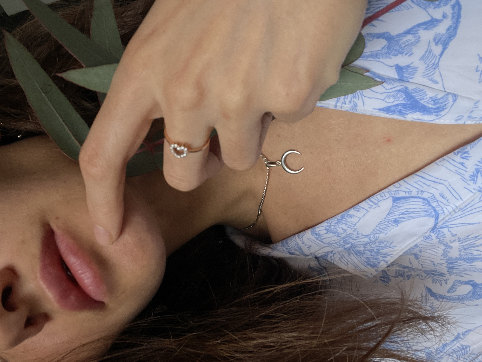 This image presents a close-up, cropped view of an individual, likely female, from the lower face to the upper chest, including a portion of the right hand. The location within Sochi, Russia, cannot be determined from the visual information provided.
The individual's lower face is visible, featuring slightly parted, full pink lips and a smooth chin area. The neck displays fair skin and a silver-colored chain necklace with a crescent moon pendant, its points facing upwards. The individual's right hand is raised, with the index finger gently touching the chin/jawline. A gold-colored ring with a heart-shaped cluster of small white stones is worn on the right ring finger. Dark brown hair, appearing long, is visible on the left side of the frame, draped over the shoulder. The individual is wearing a light-colored garment, possibly a shirt or pajama top, featuring a blue and white toile pattern depicting trees, figures, and what appear to be pastoral scenes. Several green leaves, consistent with eucalyptus foliage, are present in the upper right portion of the image, held near the person's head or positioned behind the hand. The background is dark and out of focus, providing no discernible environmental details.
