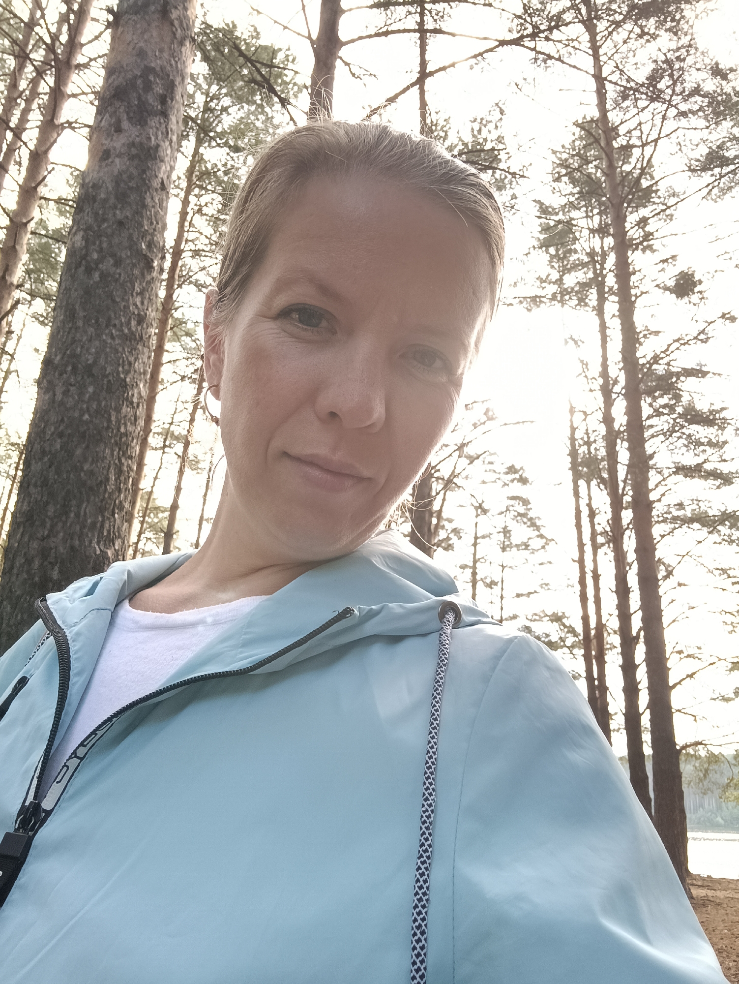 A woman is captured in a head-and-shoulder selfie, slightly angled, outdoors in a sunlit pine forest. She has light-colored hair pulled back, and her face is illuminated by soft, bright light coming from behind her. She wears a light blue or mint green waterproof jacket with a black zipper, over a white t-shirt. A black and white braided drawstring is visible on the jacket. Faint, partially visible text, possibly part of a brand name, can be seen in white letters along the jacket's zipper, reading "SYS" or "SYSTEM".
The background is dominated by tall, slender pine trees, characteristic of a northern forest, through which bright sunlight filters, creating a backlit, slightly hazy effect. The light suggests either late afternoon or early morning. On the far right, a calm body of water, likely a lake or wide river, is visible, reflecting the bright sky. The scene evokes a sense of peace and connection with nature in Chaikovsky, Russia. The woman has a subtle, pleasant expression, looking directly at the camera.