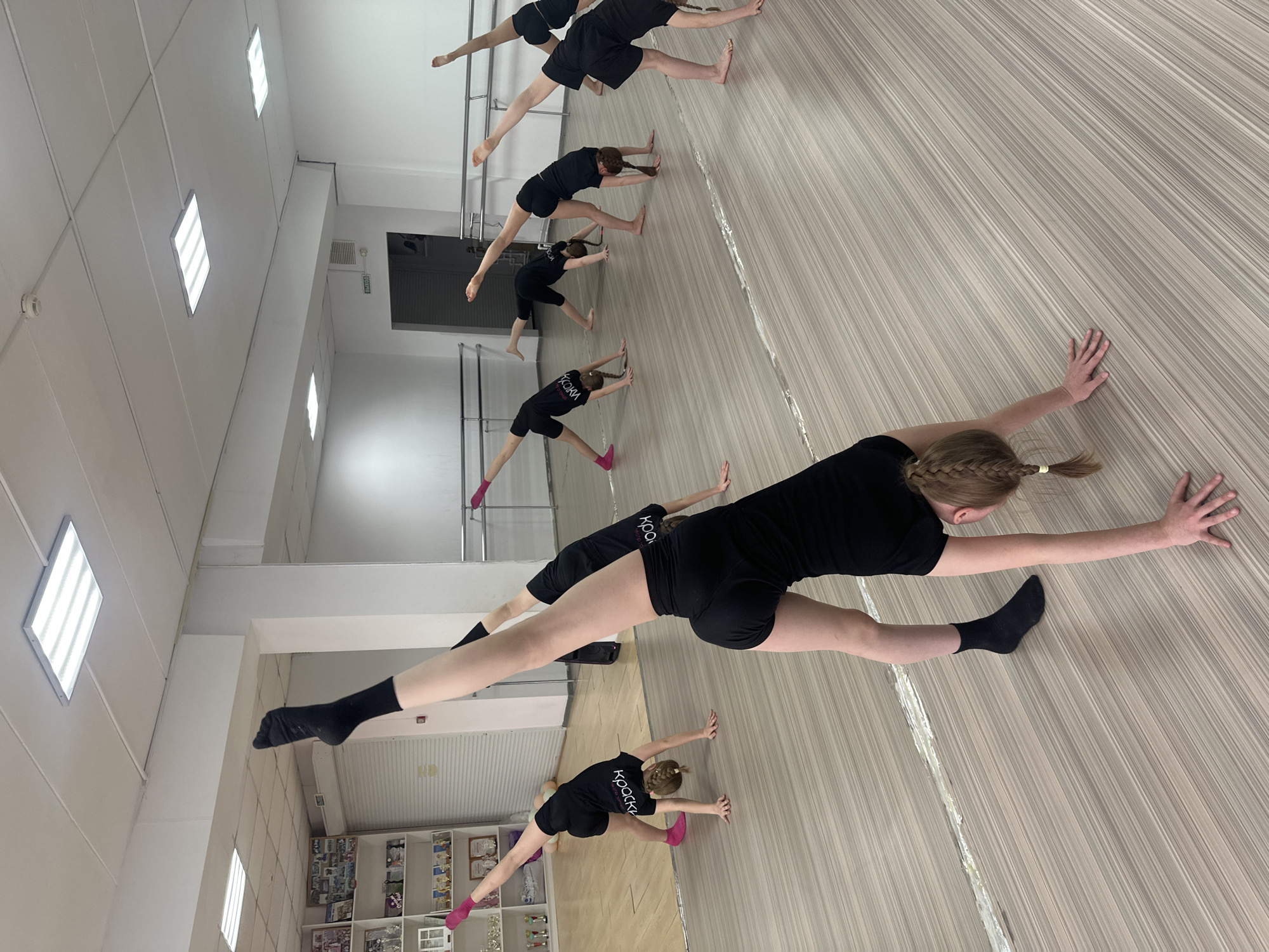 The image depicts a group of young women practicing dance stretches in a studio. They are wearing matching black shorts and tops with the word "Крошки" printed on the shirts. The women are positioned in a row, stretching with their legs apart and hands on the floor. The location is likely a dance studio, based on the ballet barre, mirrored walls, and specialized flooring. The city is Votkinsk, Russia, due to the word "Крошки" being printed on the shirts.