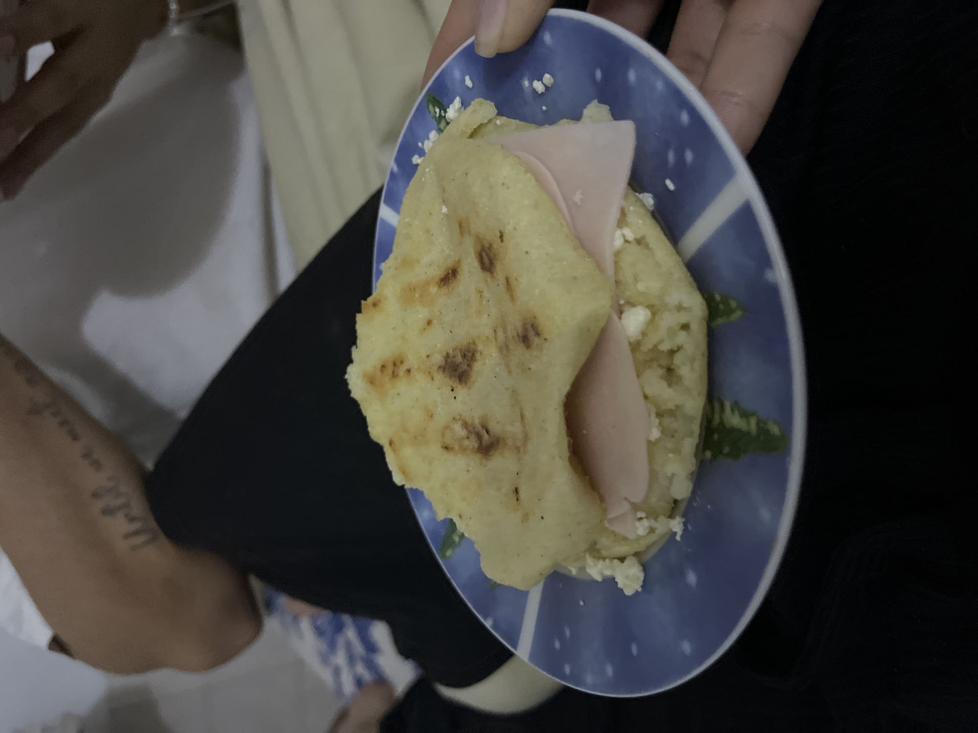 A person holds a blue plate containing an arepa filled with cheese and ham. The background shows portions of other people and furniture, suggesting an indoor setting. Juan Griego, Venezuela, is not apparent from the visual information.