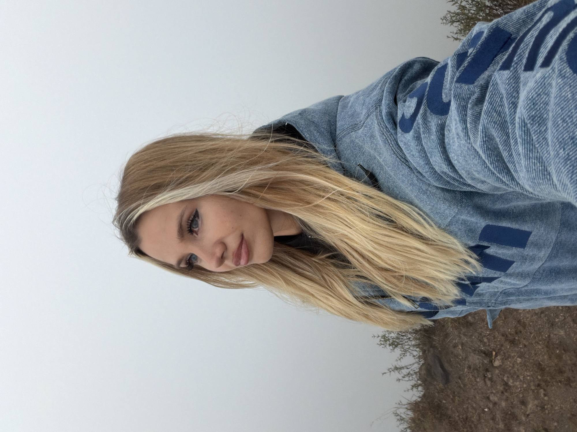 A young woman with long blonde hair is taking a selfie in the misty outskirts of Kintamani, Indonesia. She is looking to her left, with her eyes slightly open and a neutral expression on her face. Her hair is partly obscured by the hood of her denim jacket, which is adorned with large blue letters. The background is a soft gray fog, suggesting a cloudy or mountainous environment. In the foreground, the ground is covered with dry grass and dirt, indicating a natural, outdoor setting. The image appears to be taken during the day, with the natural light creating a muted color palette.