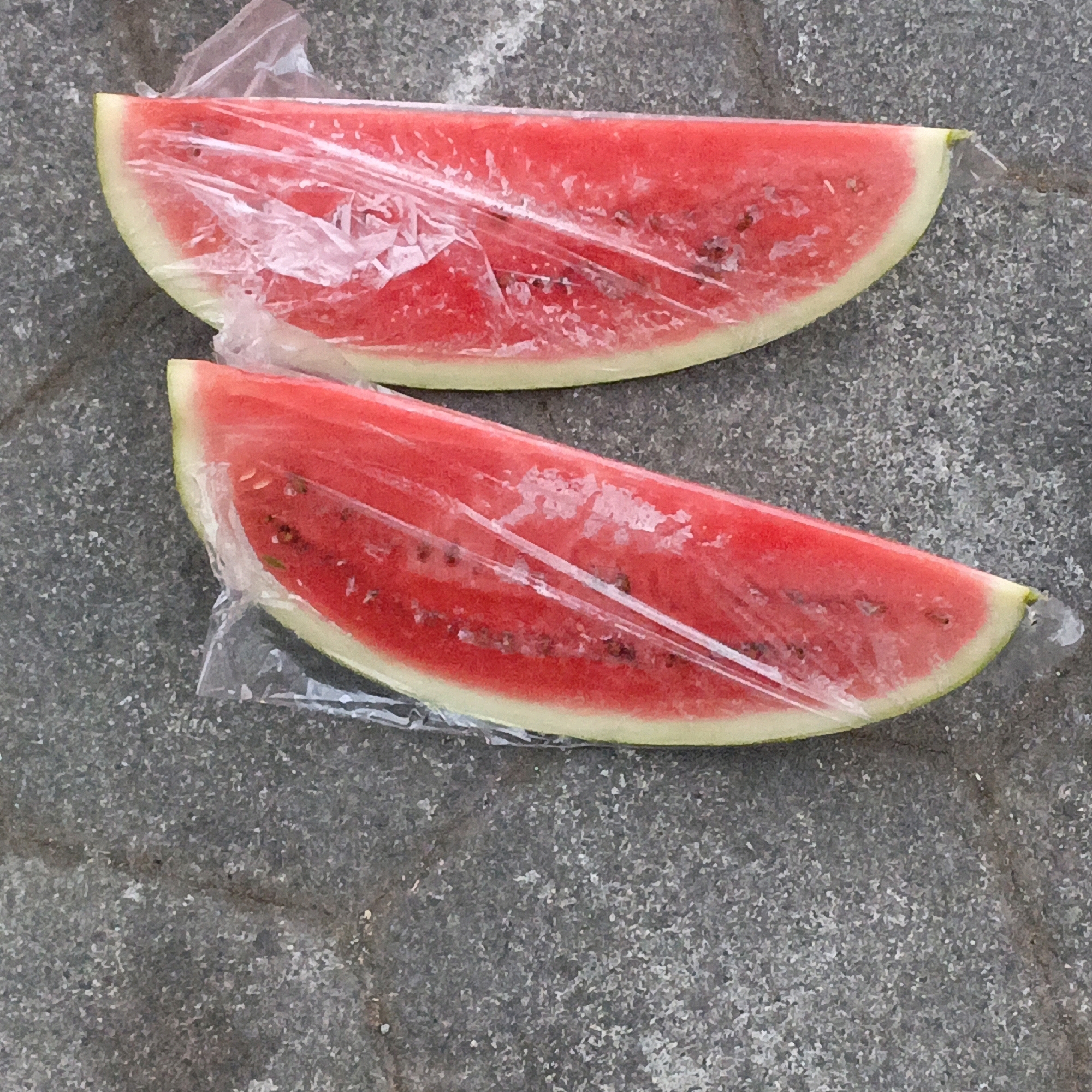 Two slices of watermelon, wrapped in clear plastic, lie on a grey, textured surface. The watermelon is bright red with visible seeds and a light green rind. The plastic wrap is slightly creased and reflects the light. The grey surface appears to be pavement or concrete, with visible small stones and variations in color and texture. The scene is likely outdoors, possibly in a public space. The location is unknown, but the image is provided in the context of Salatiga, Indonesia.