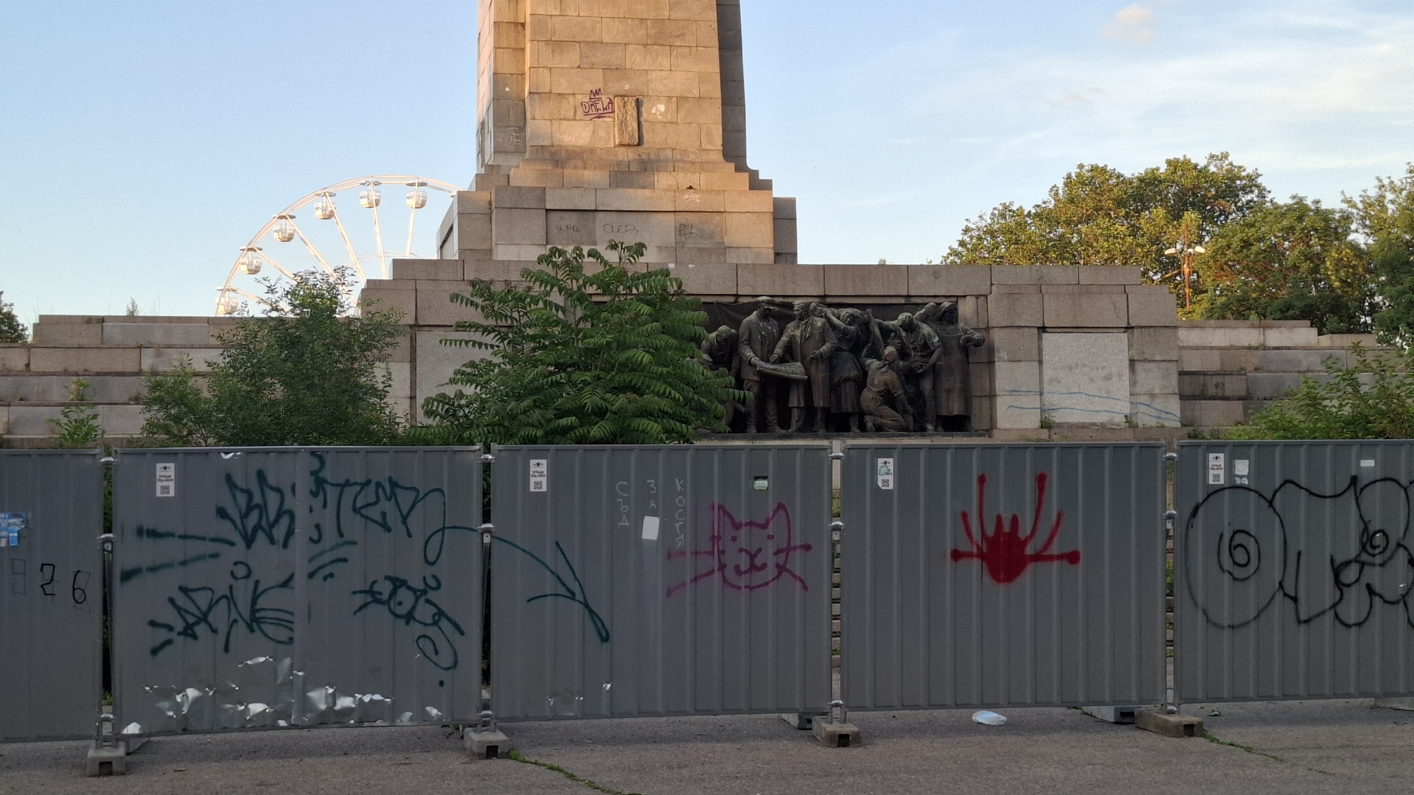 The image depicts the monument to the Soviet Army in Sofia, Bulgaria. In the foreground, a temporary metal fence with graffiti covers the base of the monument. The fence panels are grey, and various spray-painted tags and images are visible, including a cat drawing, red splatters, and black scribbles. Behind the fence, the lower section of the monument is visible, featuring a bronze relief sculpture of soldiers. A large stone pedestal rises above the relief. In the background, a Ferris wheel is partially visible against a clear blue sky, suggesting the monument is located in a public park or recreational area. Trees and foliage surround the monument, and the overall scene suggests an urban outdoor setting. The lighting indicates it is daytime, likely late afternoon or early evening, given the warm tone of the light on the monument.