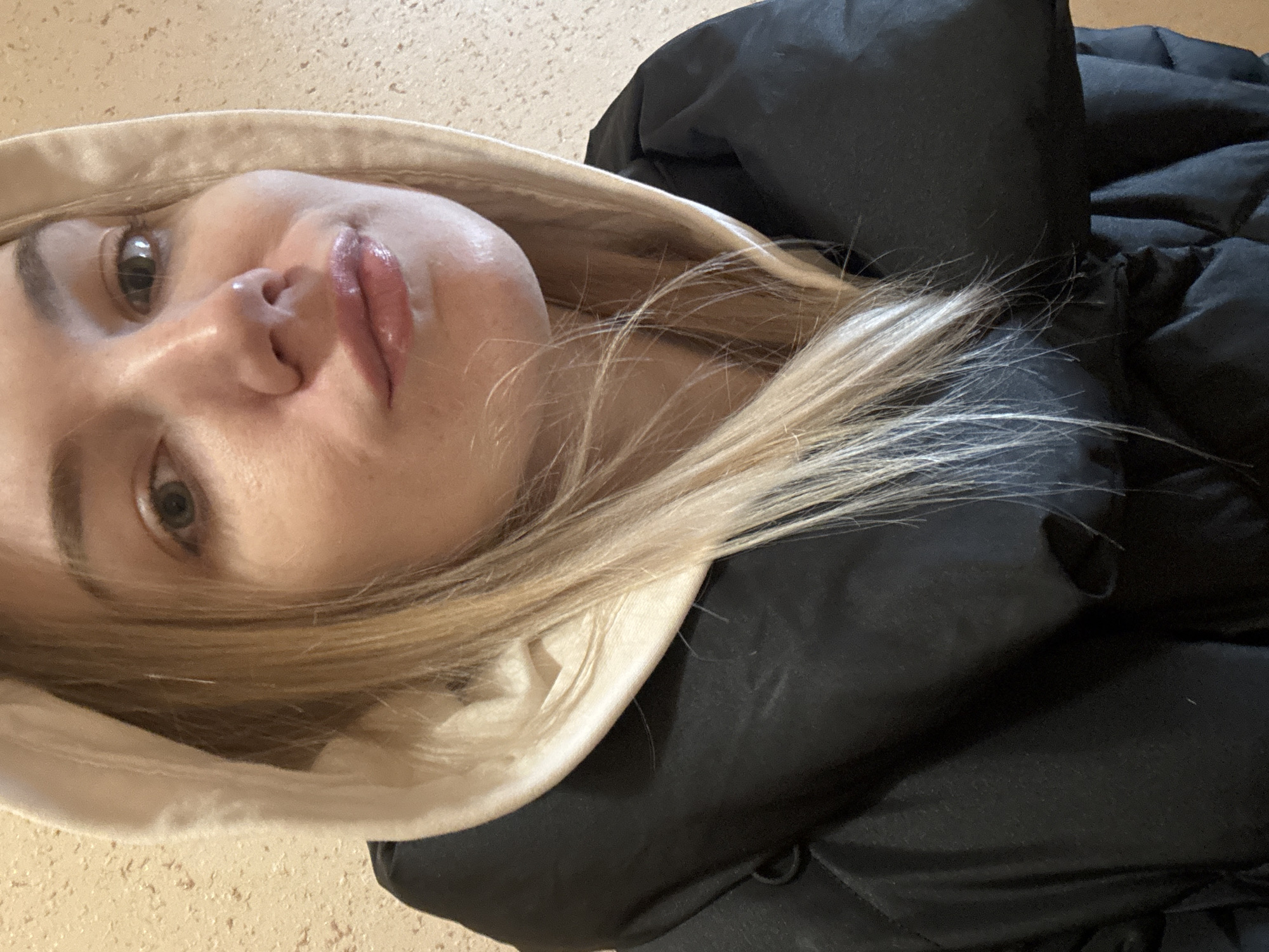 A close-up, slightly angled portrait shows a fair-skinned person with blonde hair, wearing a hooded black puffy jacket. The hood has a light-colored lining. The person has light eyes and appears to be wearing lip gloss. The background is a light tan color, possibly a floor or wall. No specific details suggest the location is Krasnodar, Russia.