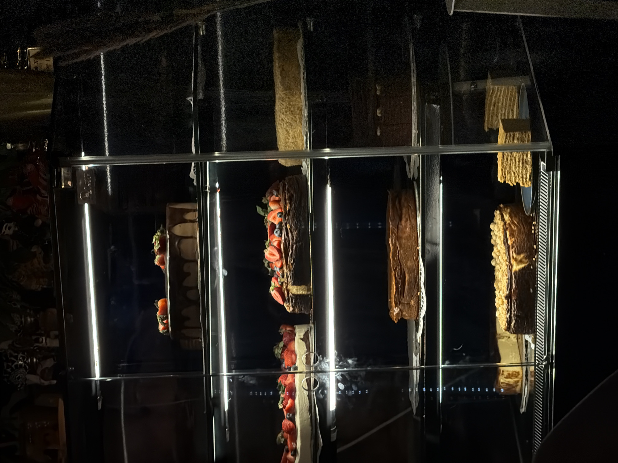 The image depicts a display case of cakes in a dimly lit environment, possibly at night. The display case has multiple tiers, each showcasing a different type of cake. The cakes include one with strawberries, one covered with a rough textured coating, and a chocolate cake. The location is likely in the city of Warsaw, Poland based on the context. The display case appears to be made of metal and glass. There are no people clearly visible in the image.