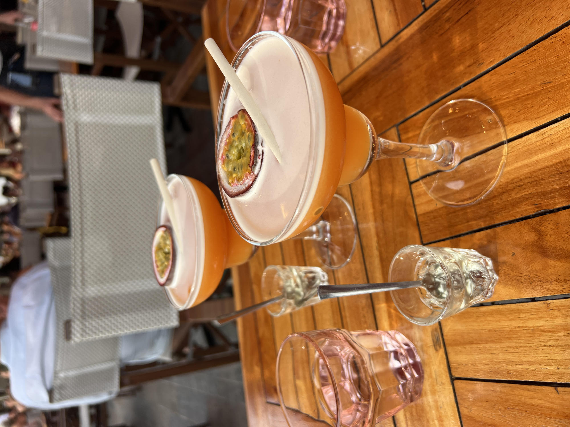 The image depicts two passion fruit cocktails on a wooden table, likely at a restaurant or bar in Cannes, France. The cocktails are served in coupe glasses, topped with a creamy foam and garnished with a slice of passion fruit and a decorative stirrer. In the foreground, there is a pink glass with a clear liquid and two small shot glasses, one containing a clear liquid and a spoon. The background shows blurred figures of people and dining chairs, suggesting a busy atmosphere. The lighting indicates it's daytime, possibly late afternoon or early evening.