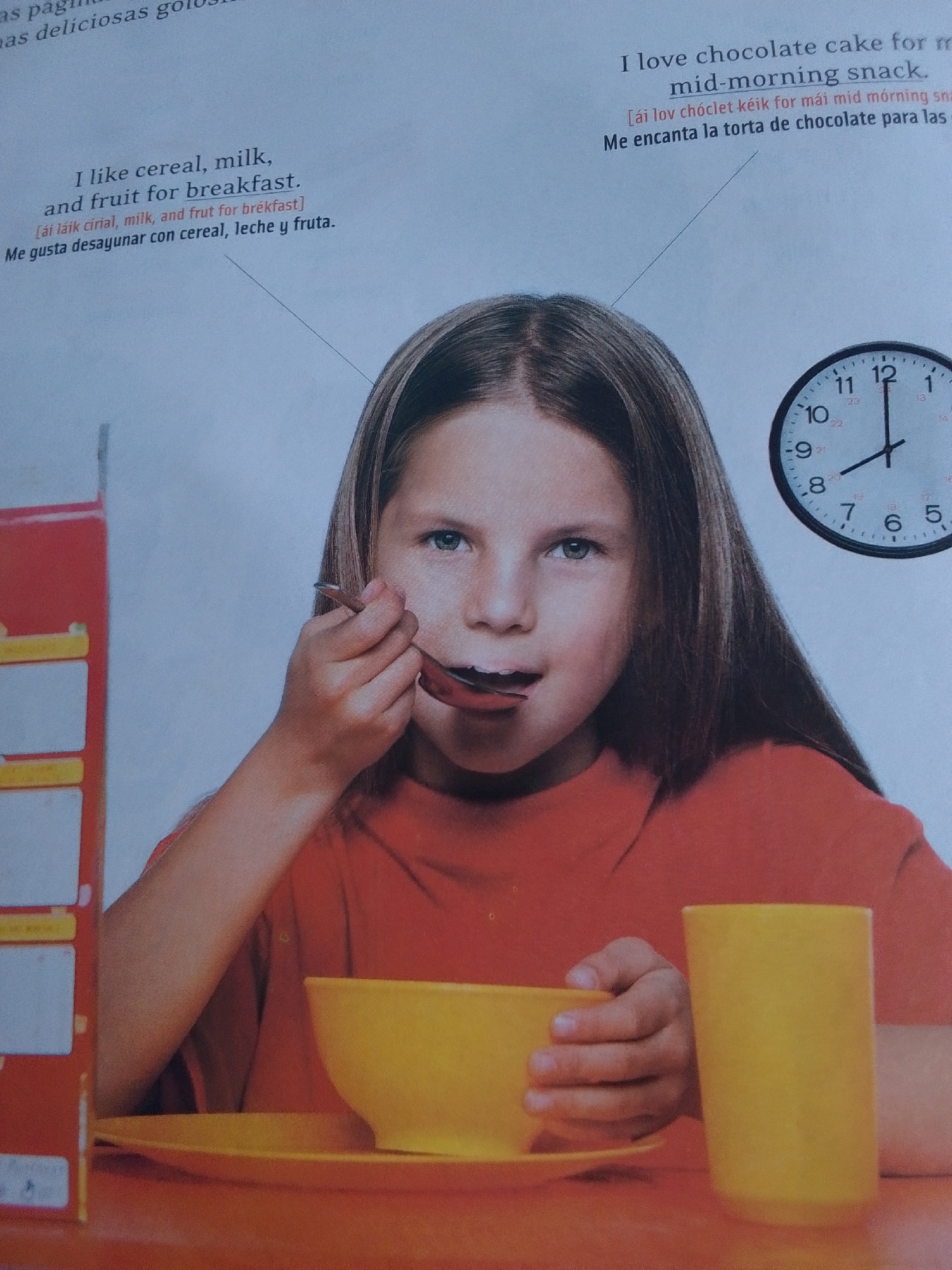 A young girl with brown hair and blue eyes is eating cereal from a yellow bowl with a spoon. She is wearing an orange shirt. A red and white cereal box is to her left, and a yellow cup is to her right. A clock on the wall behind her shows it is 11:55. The text in the image is in English and Spanish, with phonetic spellings in brackets. The provided context states the image is from the city of La Tagua, Venezuela.