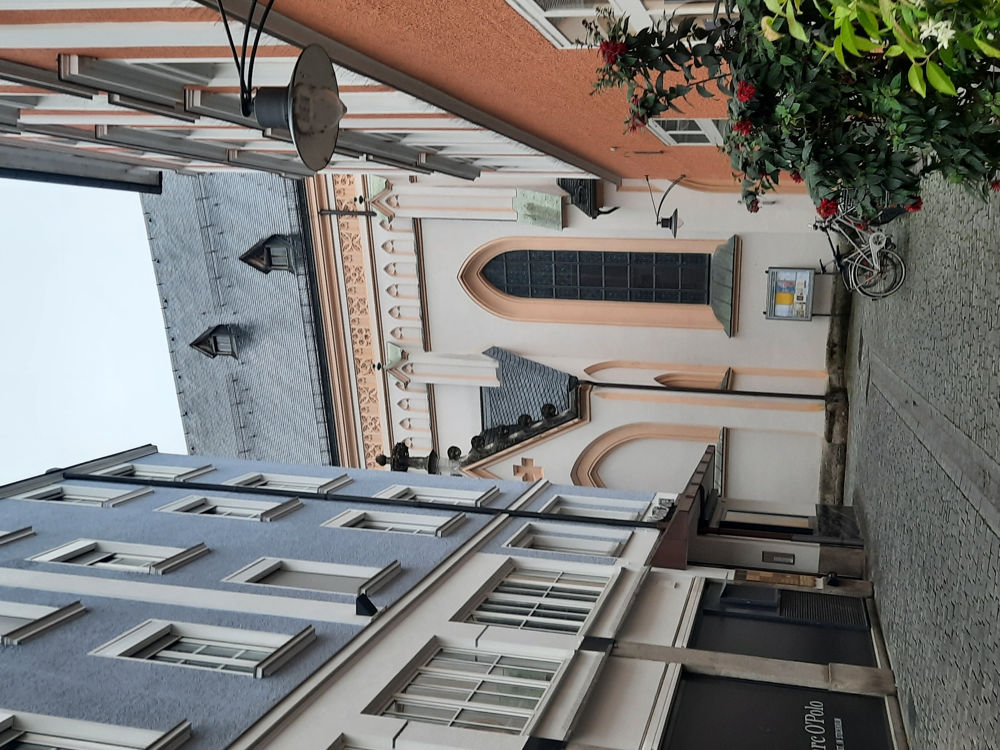 The image depicts a narrow cobblestone street in Rosenheim, Germany, lined by buildings of varying architectural styles. On the left, a light blue building with white trim features multiple windows and appears to be commercial on the ground floor, with a "Marc O'Polo" sign visible. To the right of this building, a light-colored building is visible, leading to the view of a church or religious building. This central structure has a light peach-colored facade with arched windows and decorative stone accents. The roof of this building is gray and steeply pitched, with two dormer windows visible. On the far right of the image, an orange building with white vertical stripes and numerous windows is present. A black lamp fixture is mounted on this building. In the foreground, on the cobblestone street, a bicycle is parked next to a cluster of flowering plants with red blossoms. A bulletin board is also visible near the bicycle. The sky is overcast.