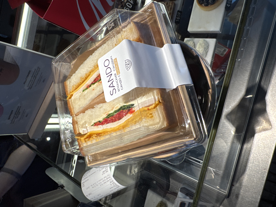 The image depicts a clear plastic container holding four triangular "Sando" sandwiches, presented in a refrigerated glass display case within a commercial establishment in Khimki, Russia. The sandwiches consist of white bread, filled with layers including yellow cheese, a red ingredient (possibly roasted pepper or tomato), green leafy vegetables, and a light-colored protein (likely chicken, as indicated by the label). A white label wrapped around the container prominently displays "SANDO" in Latin script, followed by "ЯПОНСКИЙ СЭНДВИЧ" (Japanese Sandwich) and "САНДО С КУРИЦЕЙ" (Sando with Chicken) in Cyrillic, along with a "SURF COFFEE" logo.
To the left of the sandwich container, a crumpled white paper receipt is visible on the display case's surface. In the background, other packaged dessert items are partially visible, including a small dark tart priced at "480Р" (480 Rubles). Above the display case, two menu boards with Cyrillic text and beverage images are present, indicating prices such as "450Р". A partial reflection of a person's upper body or arm is visible in the glass panels in the upper left quadrant. A red object, possibly a branded box or bag, is visible to the right. The overall setting is consistent with a cafe or coffee shop environment.