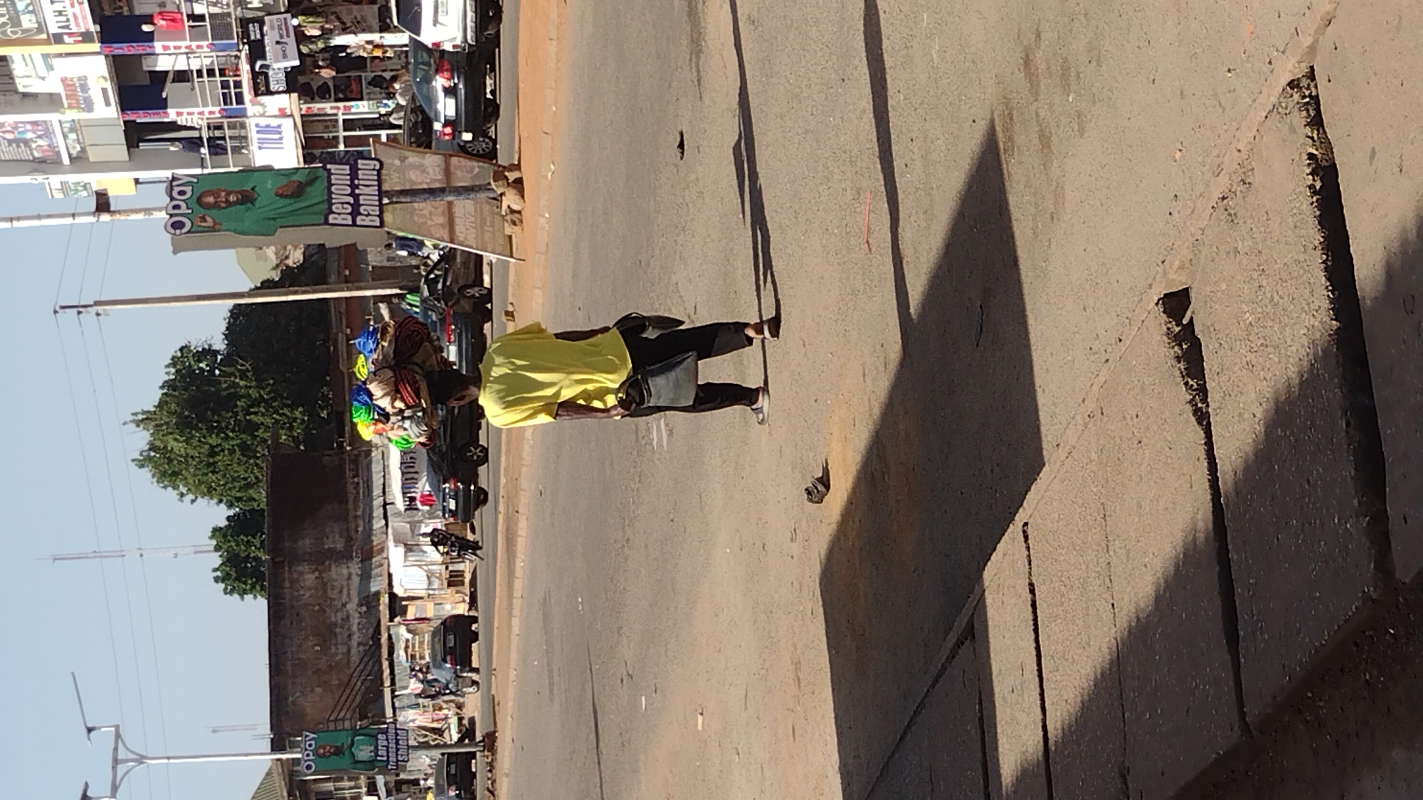 An eye-level shot captures a street scene in Jos, Nigeria. A person wearing a yellow shirt and black pants walks along the road carrying a black bag. A vendor sits next to the street, selling colorful knit caps. In the background is a billboard advertising "Opay Beyond Banking." Cars and pedestrians are present, typical of a city street. The scene is bathed in sunlight, casting long shadows on the road and sidewalk. The sidewalk is made of concrete with cracks and breaks. The sky is clear.