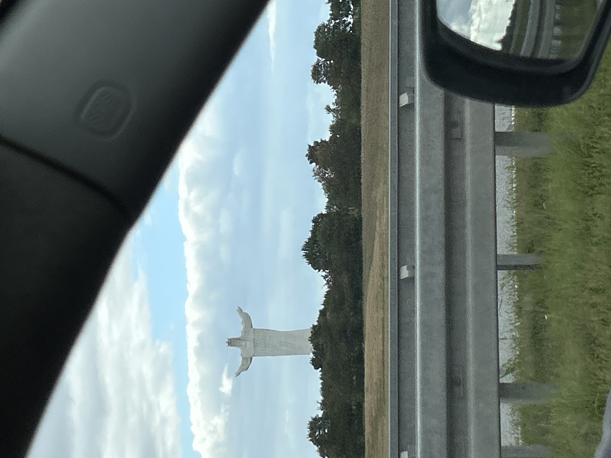 The image shows a large white statue of Jesus Christ with outstretched arms, wearing a crown, situated on top of a hill surrounded by trees. The statue is the Christ the King statue in Świebodzin, Poland, which is often mistaken for being located in Lubinicko. The statue is visible in the background, seen from a vehicle's perspective. In the foreground, there is a highway guardrail, and the reflection of the sky and trees can be seen in the side mirror of the vehicle. The sky is partly cloudy.