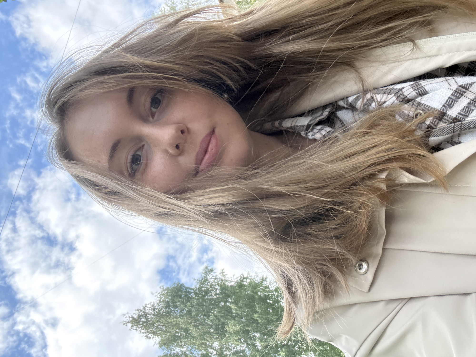 A young woman with fair skin and light brown hair poses for a selfie outdoors. The background includes a sky with scattered clouds and a leafy green tree. She is wearing a light-colored jacket with a button visible. Under the jacket, a checkered pattern can be seen. The location is Unknown, Unknown.