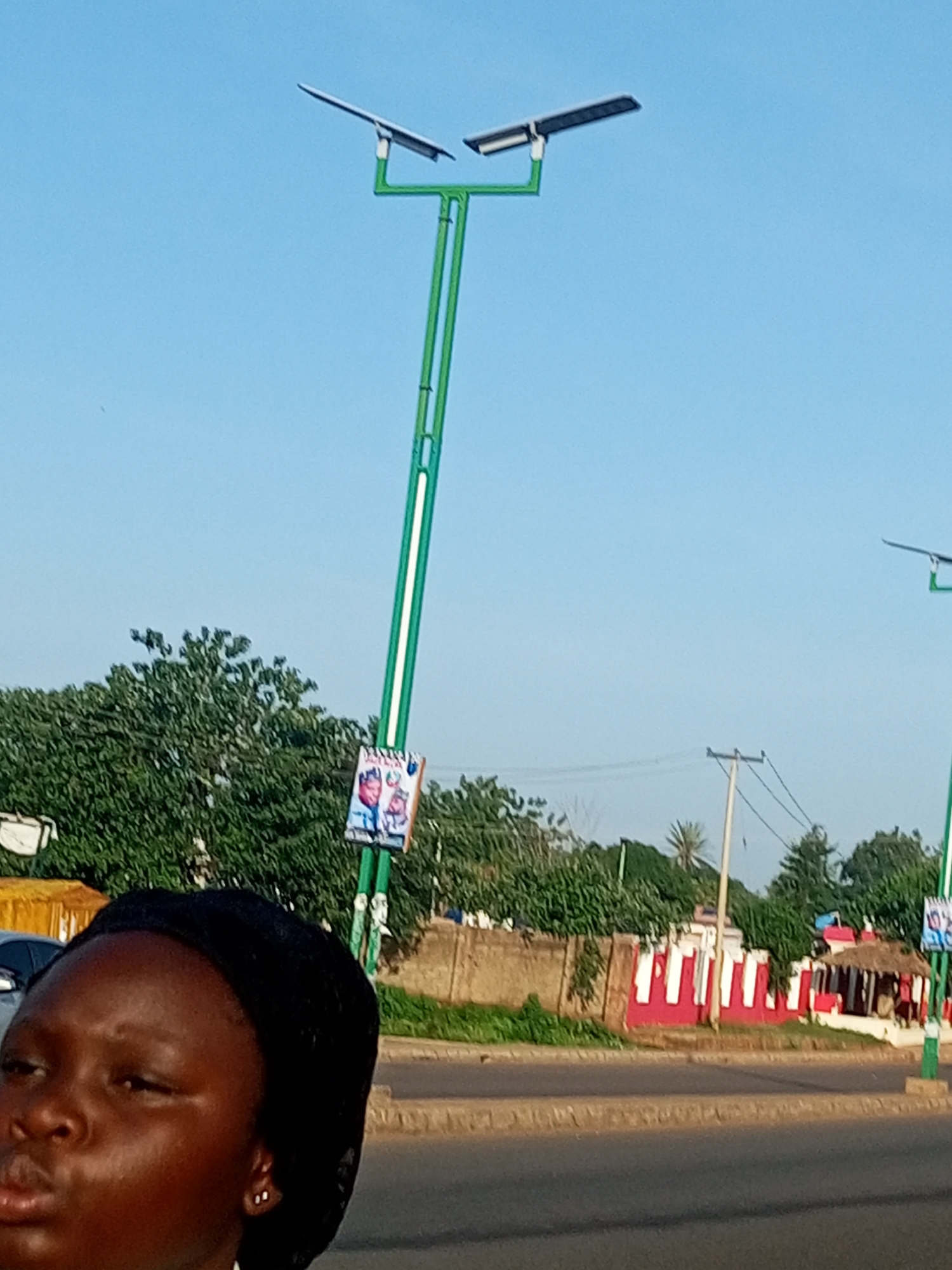 A close-up shot focuses on the left side of a young Black girl's face, who is wearing a black headscarf and small white earrings. Her expression is neutral, with her gaze directed to the left. Behind her, the scene transitions to a street in Garko, Nigeria. A two-pronged green lamppost with solar panels is prominently featured, with a small poster affixed to its base. The poster depicts two individuals, one in red and white, and another in blue and white. Beyond the lamppost, a dense cluster of green trees forms a backdrop. Further in the distance, a beige wall with a series of alternating red and white vertical stripes runs along the street. A small thatched-roof structure with red accents is visible behind the wall. A power line pole with wires stretches across the middle ground. The sky is a clear, light blue.