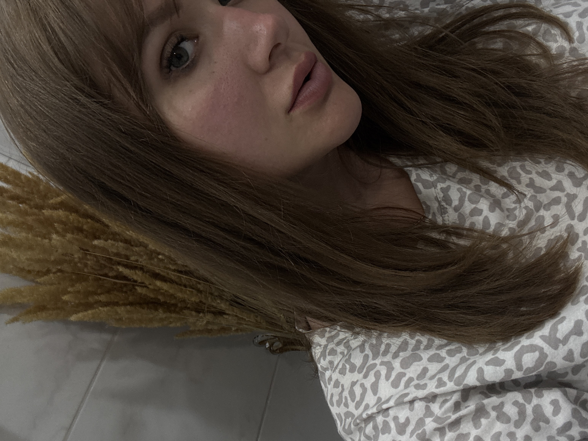 The image shows a woman with light brown hair and fair skin lying down. Her face is angled towards the camera, revealing her eyes, nose, and lips. She is wearing a light-colored top with a grey animal print. Beside her is a bundle of dried wheat or grass. The background appears to be a light-colored tiled floor. There is no indication that the photo was taken in Stavropol, Russia.
