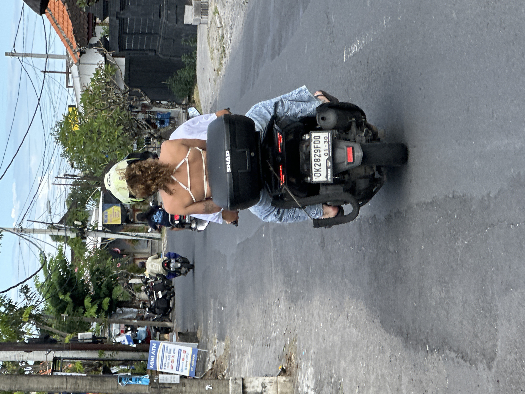 The image shows a street scene in North Kuta, Indonesia. A woman with curly hair, wearing a white top with spaghetti straps and blue patterned pants, is riding a motorcycle. She is wearing a pale yellow helmet. The motorcycle has a black storage box on the back. The license plate reads "DK2829FDQ" with the date "01-30". The road is paved and shows signs of wear. Several other motorcycles are visible in the background. There is a "MINI ATM" sign beside the street. Buildings, trees, and power lines line the street.