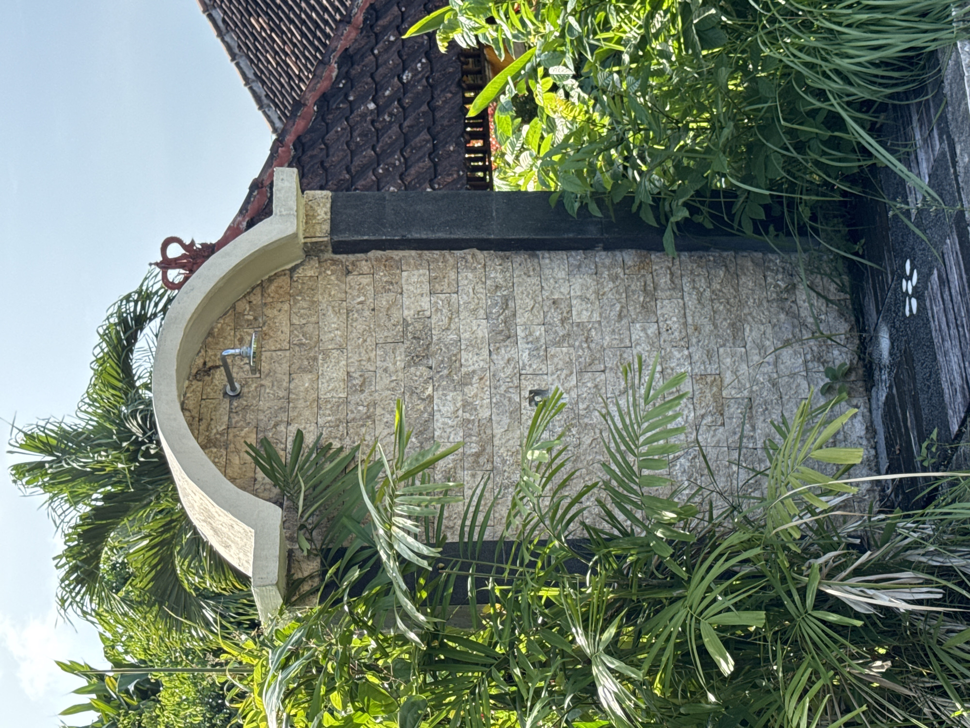 A sunlit, outdoor shower is surrounded by lush greenery. The shower area is constructed with a curved wall of tan brick tiles, and a gray, textured stone border runs along the top of the shower wall. A metal shower head is mounted on the brick wall. A wooden surface with white decorative inlays forms one side of the shower base. The shower is set against a backdrop of a tiled roof and tropical vegetation. The location is Ungasan, Indonesia. No people are visible.