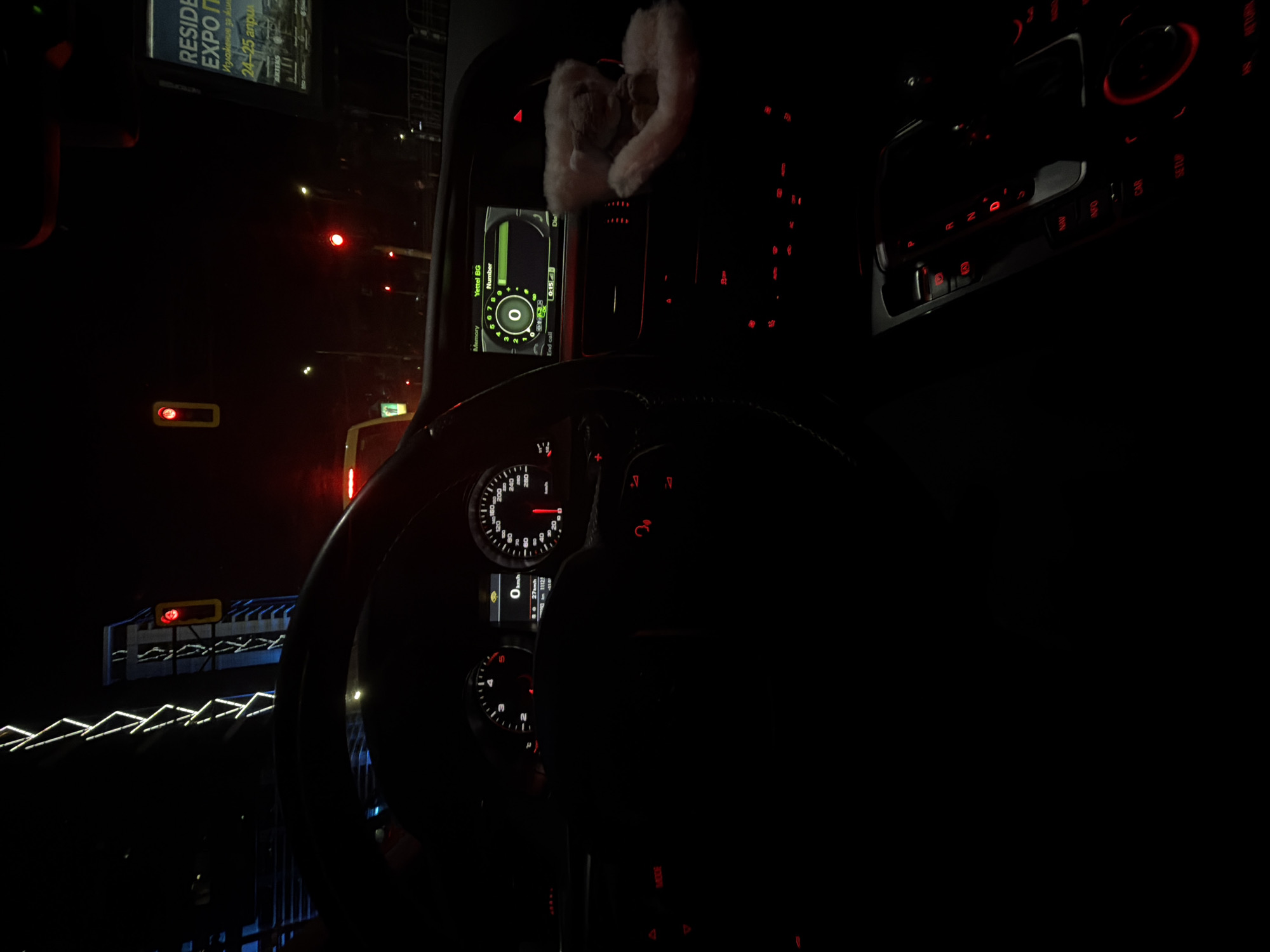 Here's a description of the image:
**General Description:**
The image shows a nighttime view from inside a car in what appears to be Sofia, Bulgaria. The scene is dimly lit, primarily by dashboard lights and faint external city lights. The primary focus is on the car's interior, particularly the steering wheel and the dashboard instruments.
**Environment and Location:**
The car is situated near a fence. Outside the car window, there are traffic signals and streetlights. A billboard that reads "Residence Expo" in Cyrillic script is faintly visible, further corroborating the location as Bulgaria.
**Objects:**
* **Car Interior:** Steering wheel, dashboard instruments (including speedometer and navigation screen), gear shift area, and car infotainment system.
* **Dashboard Decorations:** There's a fuzzy ornament hanging from the rearview mirror.
* **Exterior:** Traffic lights, streetlights, a metal fence and a billboard
**Details from the Dashboard:**
* The speedometer displays a speed of approximately 27 km/h.
* The navigation system is active, with a call in progress for 0:15 seconds.
* The network provider "Yettel BG" is displayed.
**Action:**
The car seems to be either moving slowly or stopped, given the speed shown on the speedometer and the presence of visible traffic signals.