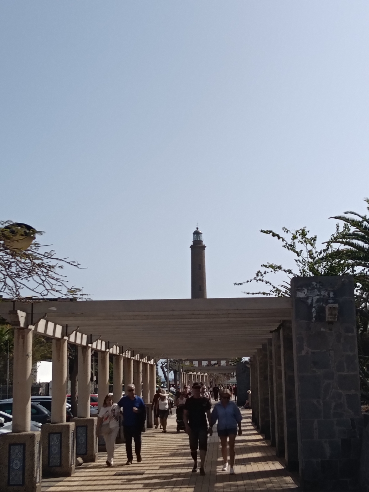 The image shows a walkway in Maspalomas, Spain, with a pergola providing shade over a pedestrian pathway. People are walking in both directions along the walkway. The Maspalomas Lighthouse is visible in the distance, centered in the frame. The sky is clear and bright blue. Trees and vegetation flank the walkway on both sides. Cars are parked on the left side of the walkway. The pergola is constructed of light-colored columns and beams. The walkway is paved with light-colored paving stones.