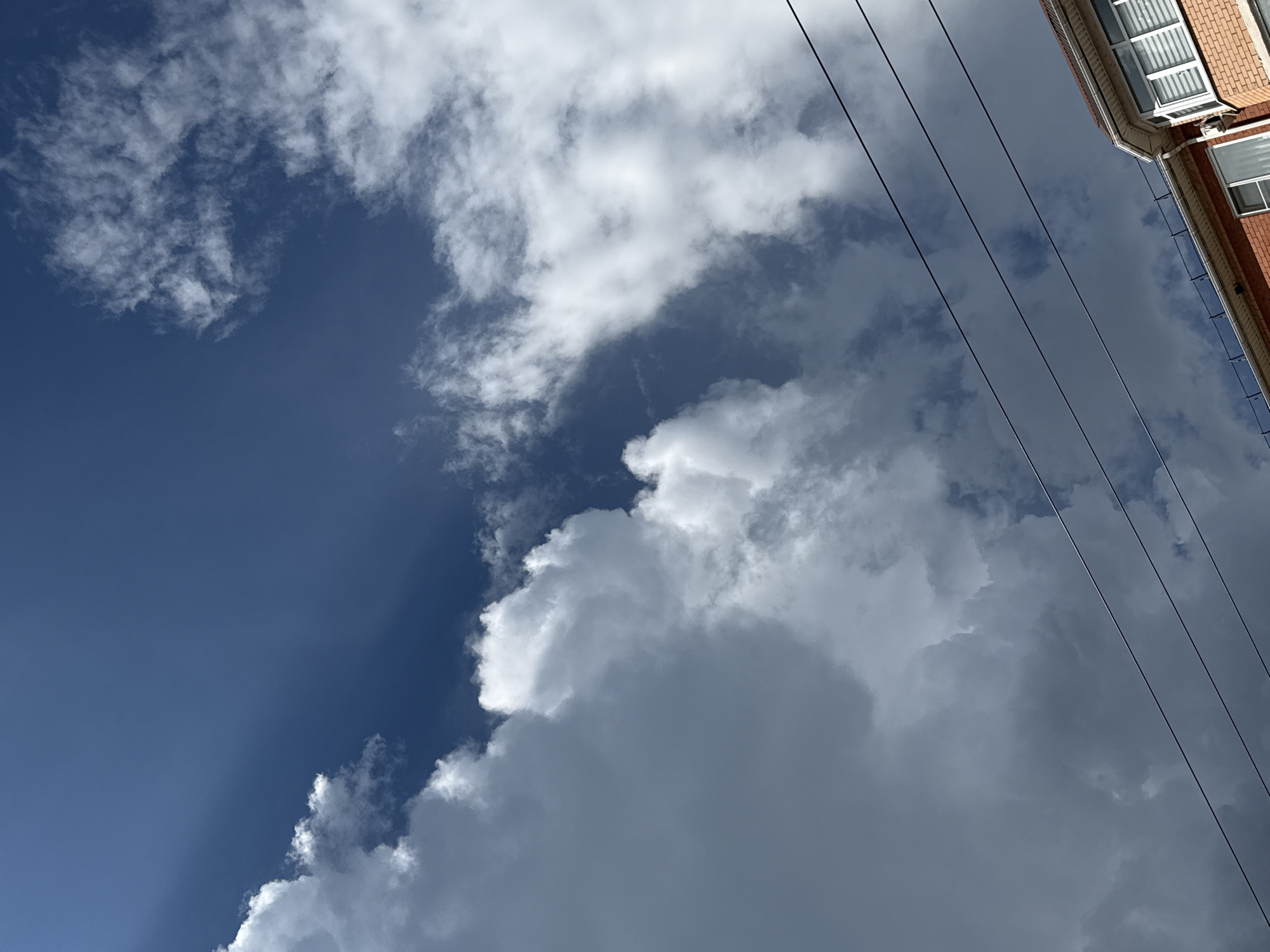A low-angle shot reveals a partially cloudy sky over an urban environment. The sky transitions from a clear, deep blue in the lower left to scattered, puffy white clouds at the top. The right side of the frame is dominated by denser, grey storm clouds. Overlapping the upper-right corner is the corner of a brick building, with white-framed windows visible. Several power lines stretch diagonally across the image, originating from the building and extending into the sky. The location is likely Unknown, Unknown.