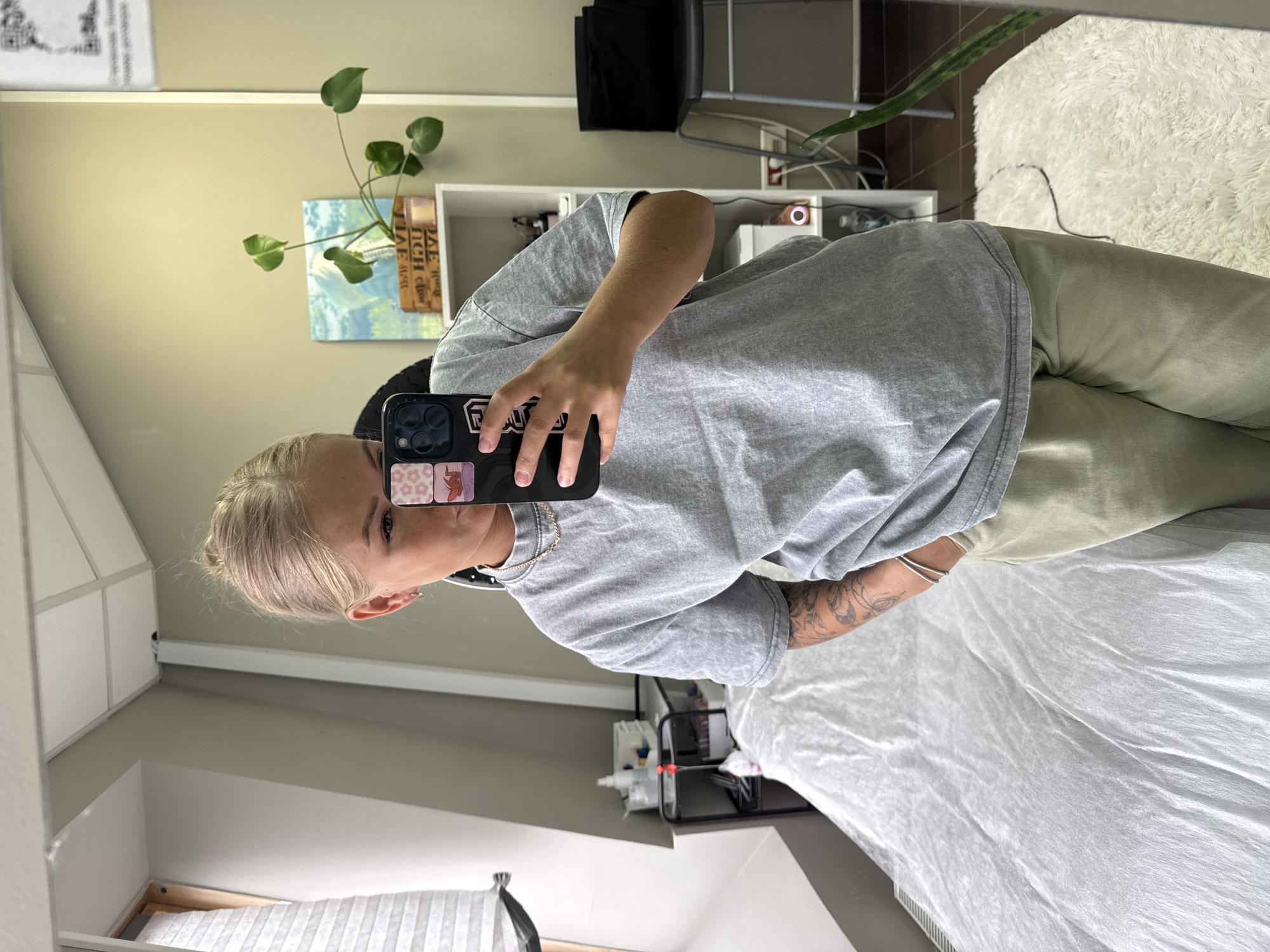 A mirror selfie taken indoors. A woman with blonde hair is holding a phone, presumably to take the photo. She is wearing a grey t-shirt and green pants. A chair and white shelf with items on it are visible in the background, along with a plant. A bed with white sheets is also visible to the right. The location is likely a home or apartment interior.