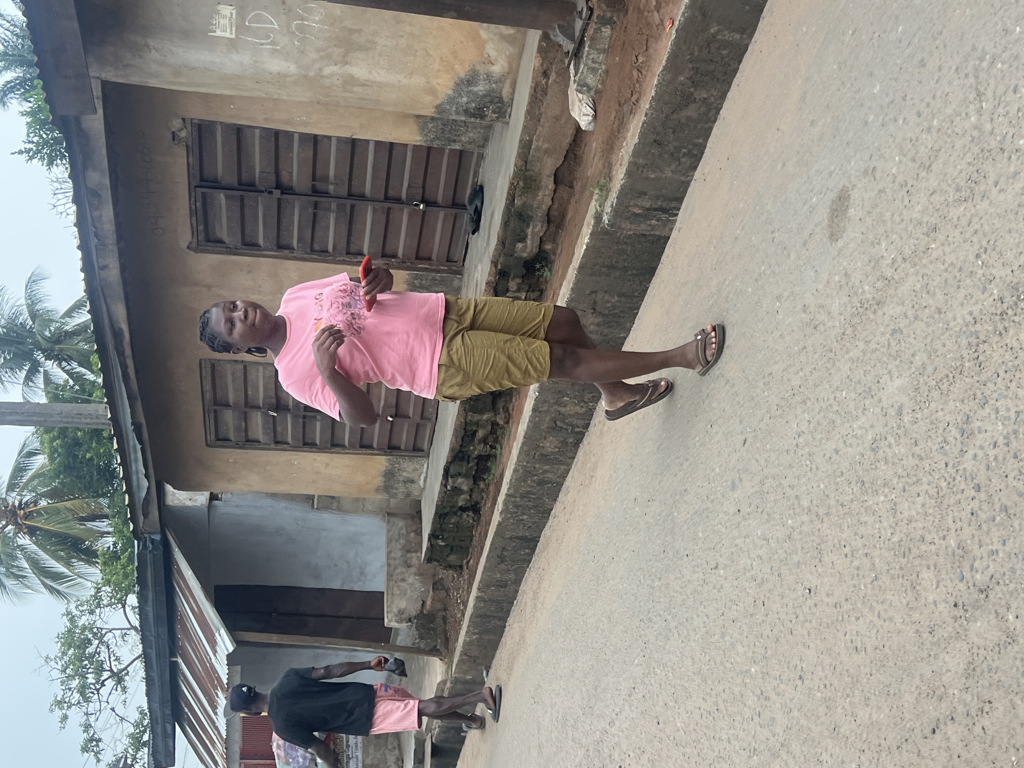 The image depicts a person standing on a paved road next to a building. The individual wears a pink t-shirt and tan shorts, holding a red object. They are wearing sandals. Another individual is seen in the distance wearing a dark shirt and pink shorts with a black baseball cap.
The building adjacent to the road shows signs of weathering and has a corrugated roof and a rusted roof. There are barred windows. A palm tree is visible in the background.
Based on the image, the location appears to be Obosi, Nigeria. The surrounding area seems to be residential.