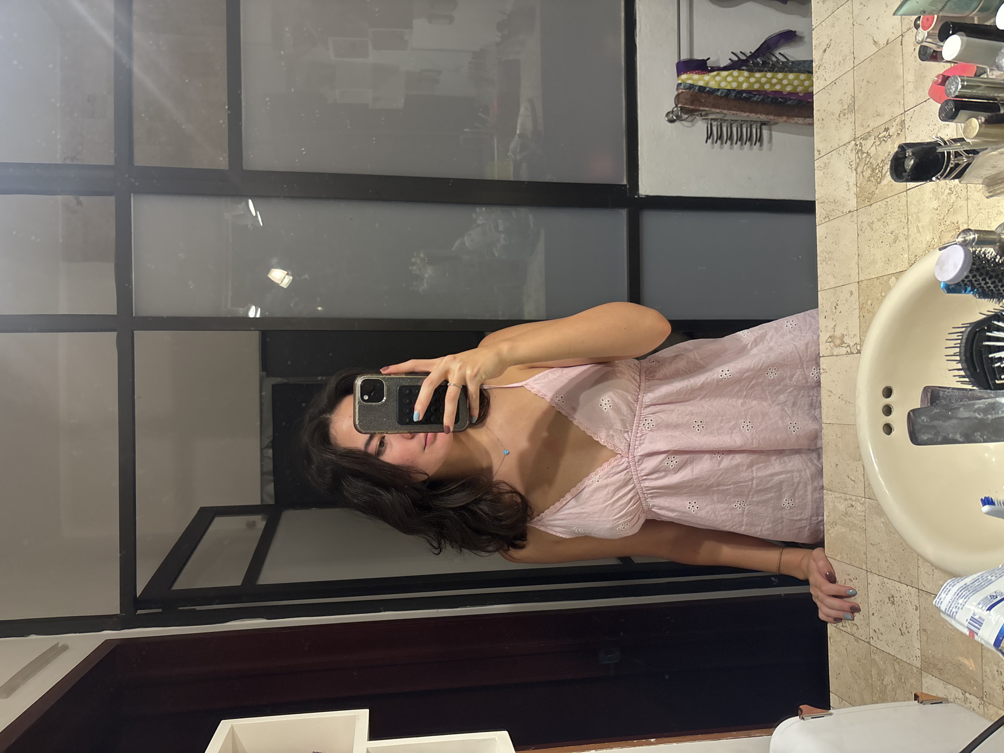 A woman is taking a selfie in a bathroom. She is wearing a light pink dress with white detailing. Her dark hair is loose around her shoulders. She is holding a cell phone with her left hand. The background shows a sink, counter with toiletries, and a window or mirror with a dark frame. The location is Puebla, Mexico.